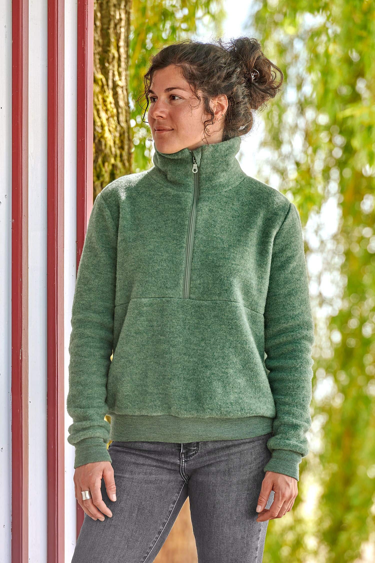 Frau steht neben rot weißem Haus und trägt grünen Wollfleecepulli aus bio Wolle (kbT) vom Finkhof