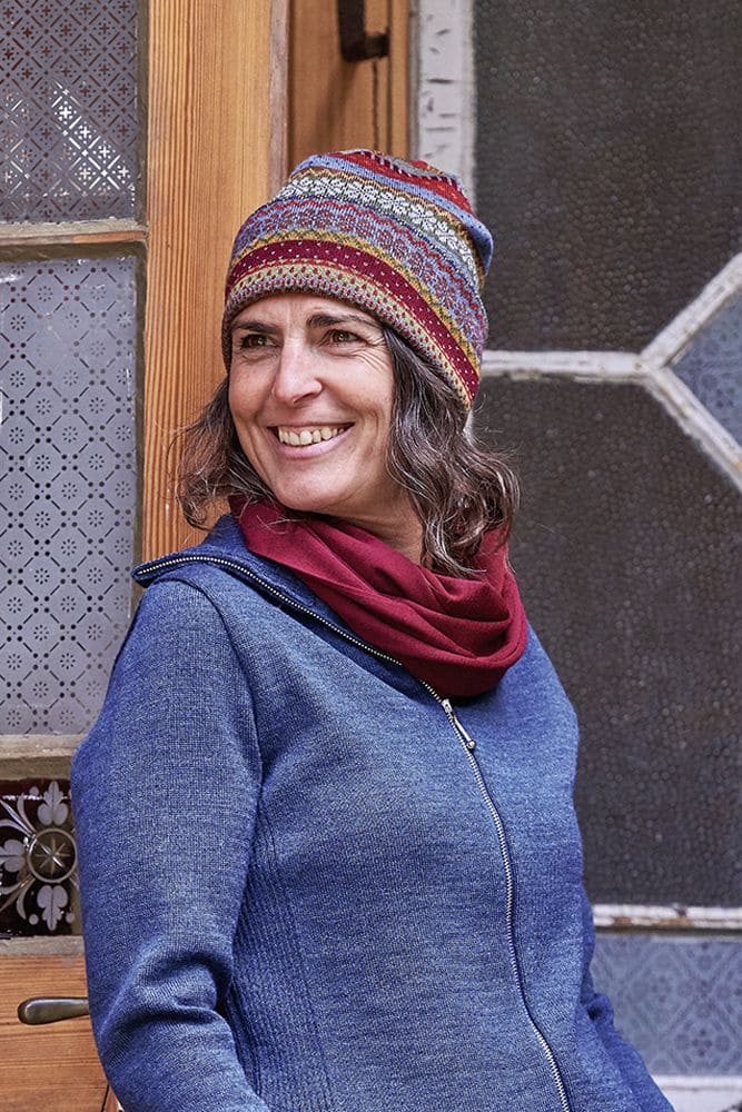Frau trägt gemusterte Mütze, roter Schal und blaue Zipperweste. Sie steht vor einer Holztür und lacht