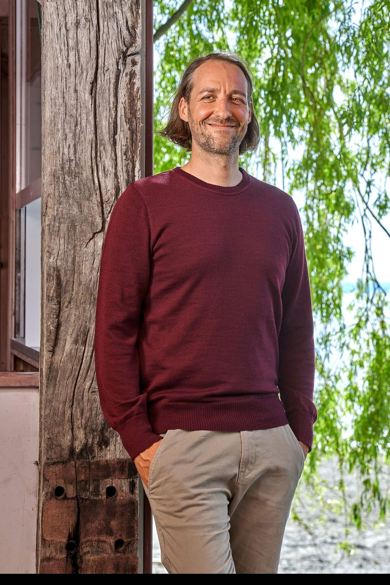 Mann mit feinem burgunderroten Wollpullover aus bio Merinowolle (kbT) vom Finkhof lehnt an einem Balken