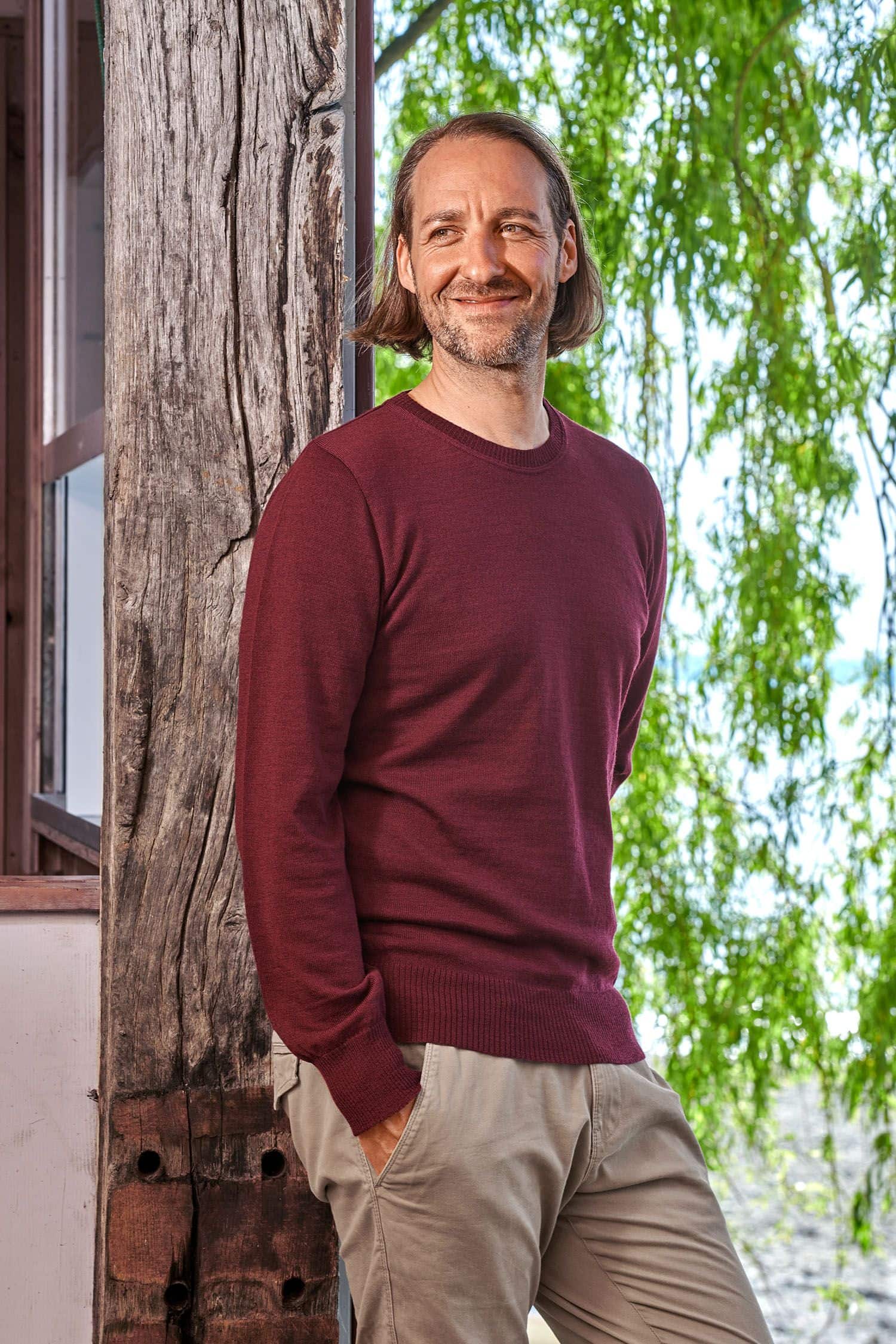 Mann mit feinem burgunderroten Wollpullover aus bio Merinowolle (kbT) vom Finkhof lehnt an einem Balken