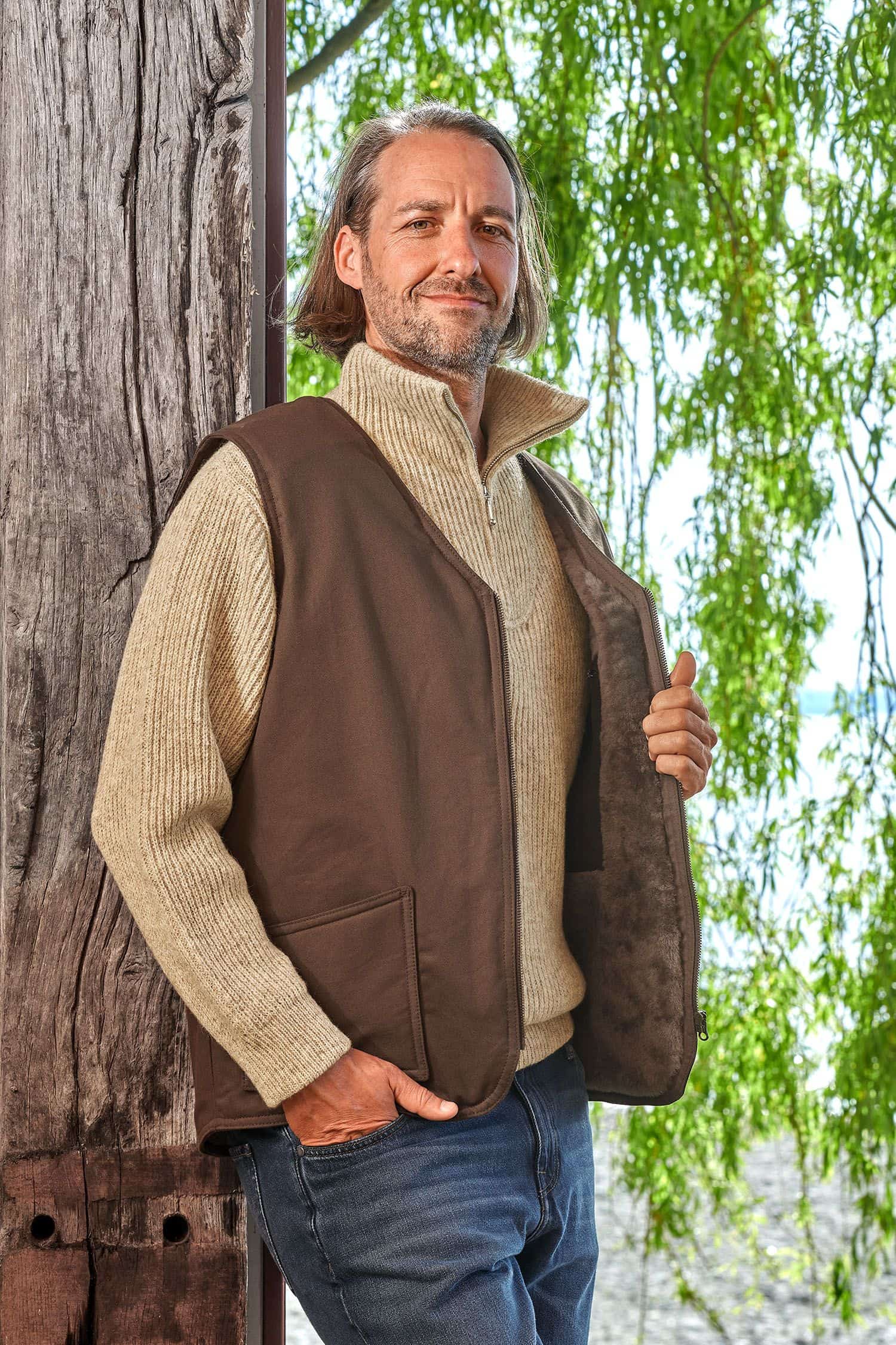 Mann lehnt sich an Baum und trägt einen Troyer und die braune Fellweste aus bio Baumwolle (kbA) und Fell vom Finkhof