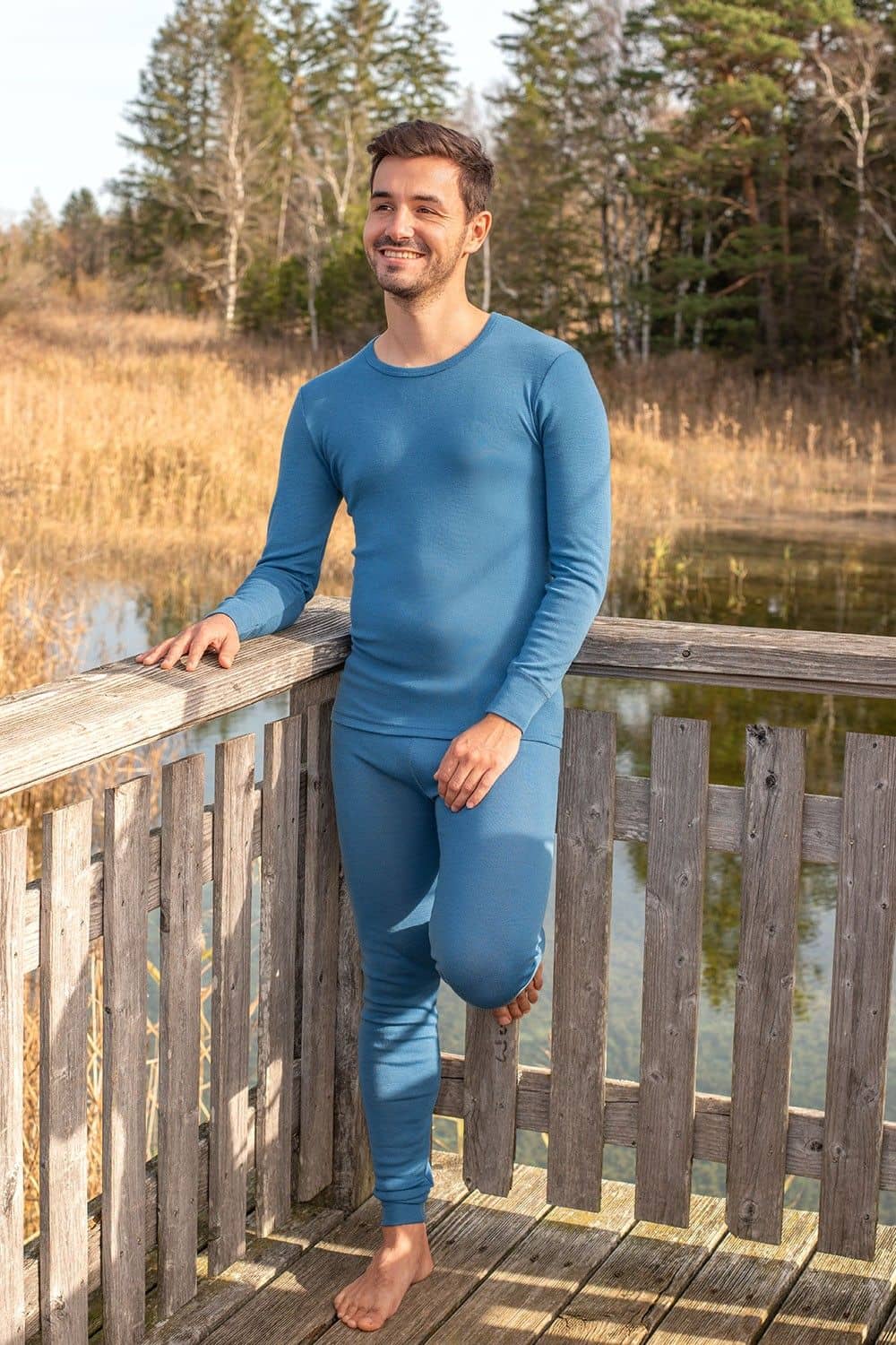 Mann  lehnt an Geländer und trägt blaue lange Unterwäsche aus bio Wolle (kbT) mit Shirt und Hose