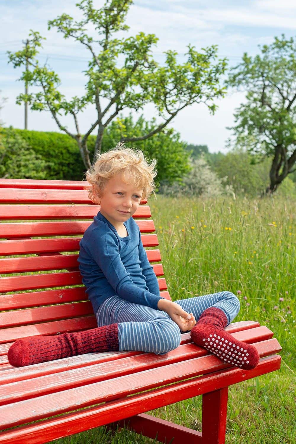 Kind sitzt auf roter Bank und trägt blaue Kleidung mit roten Wollplüsch Stoppersocken aus bio Wolle (kbT)