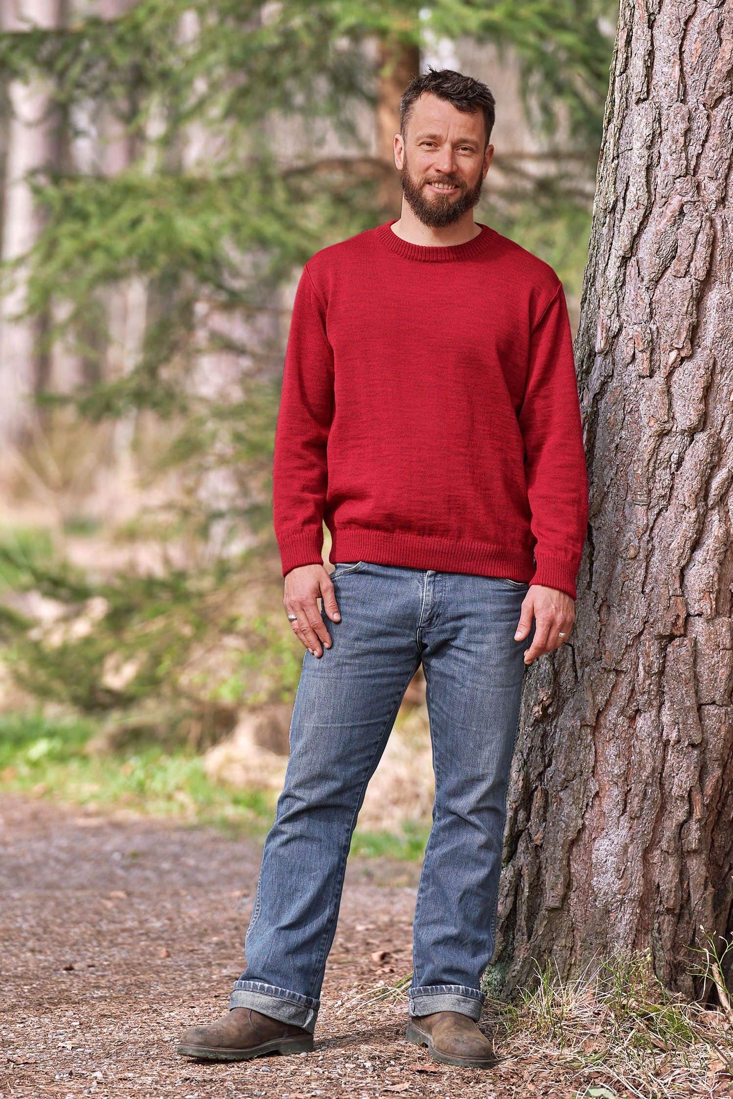 Mann steht mit Jeans und rotem Rundhalspullover aus bio Wolle (kbT) vom Finkhof neben einem Baum