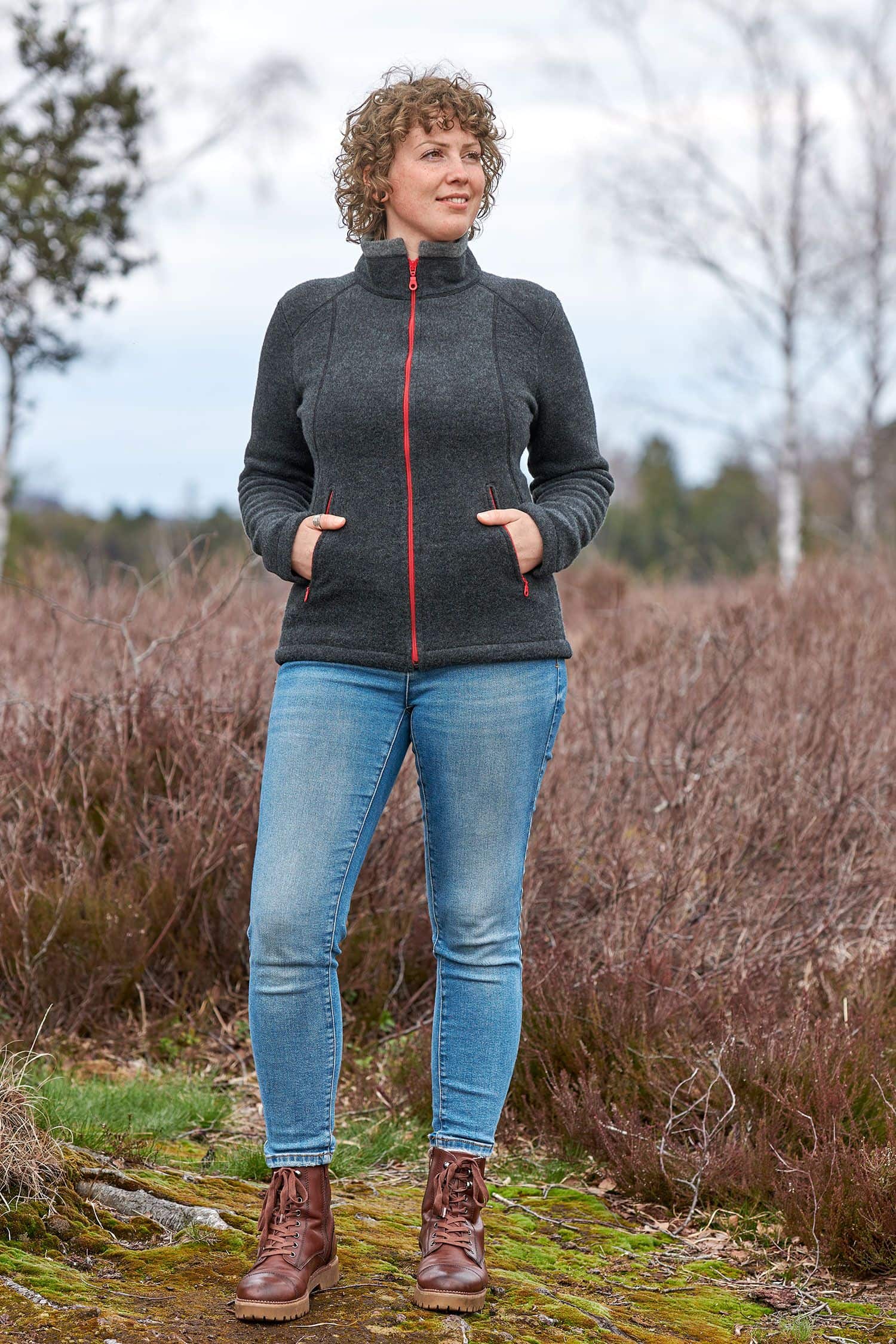 Frau mit Jeans trägt eine anthrazitgraue Wollfleecejacke mit Stehkragen und roten Reißverschlüssen aus bio Wolle (kbT)