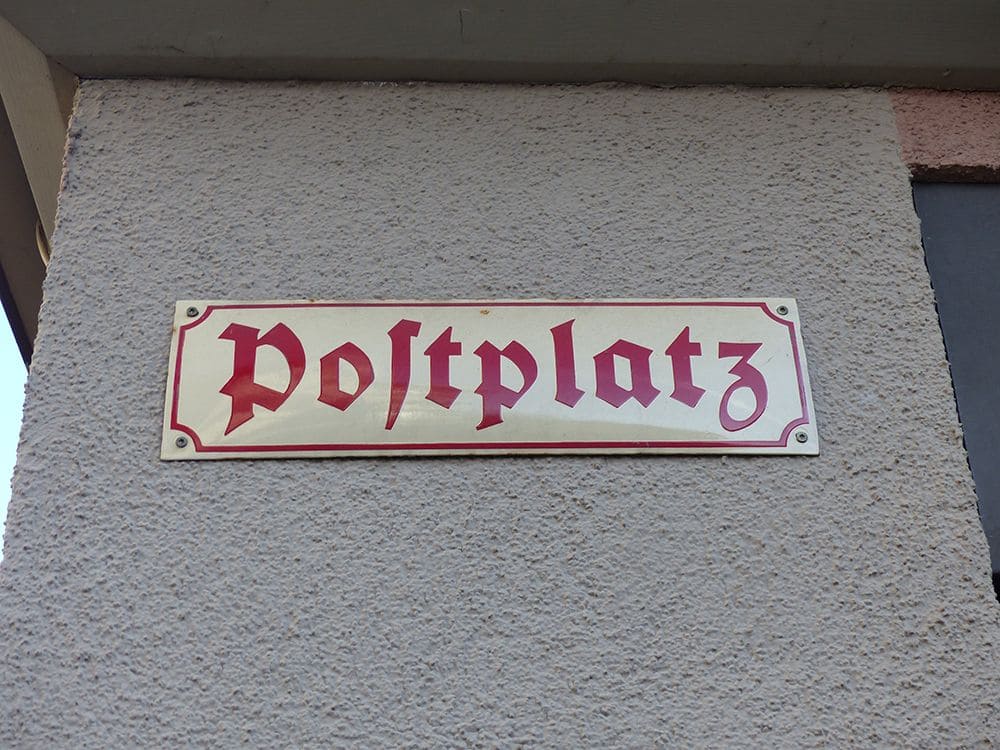 Schild_Postplatz weißes Dorfplatz Schild mit roter Schrift an grauer Wand