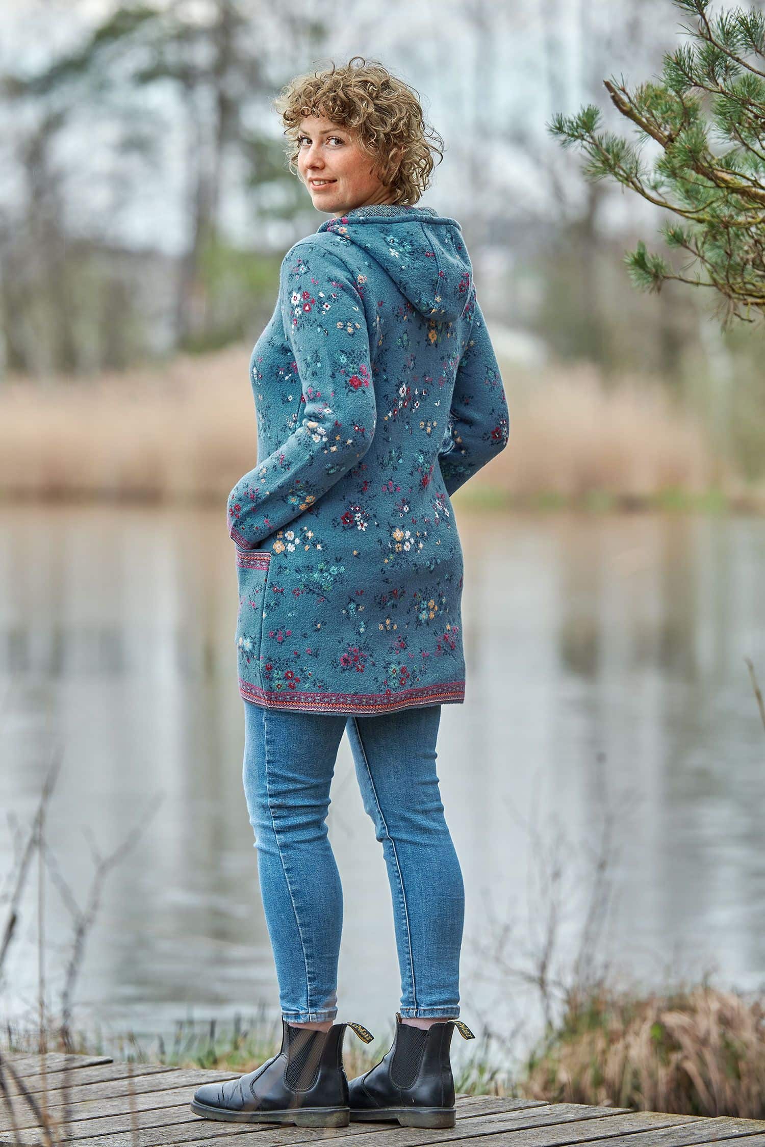 Walk-Kapuzenjacke Nelia stahlblau Frau auf Steg trägt lange blaue Kapuzenjacke aus Walkstoff mit bio Merinowolle (kbT) und Baumwolle (kbA) mit Blumenmuster