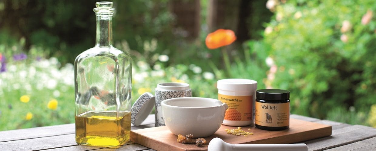 Ölflasche, Bienenwachs, Wollfett, Blüten und Mörser auf einem Tischchen im Grünen
