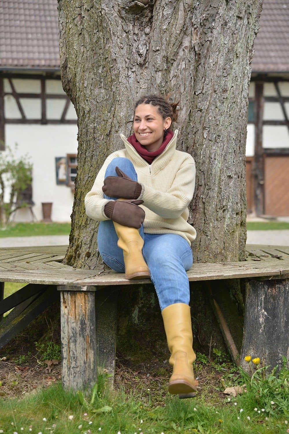 Frau sitzt auf Bank und trägt dunkelbraune Fell- Fausthandschuhe aus Pelzvelour mit Bund, weißer Pulli und roter Schal