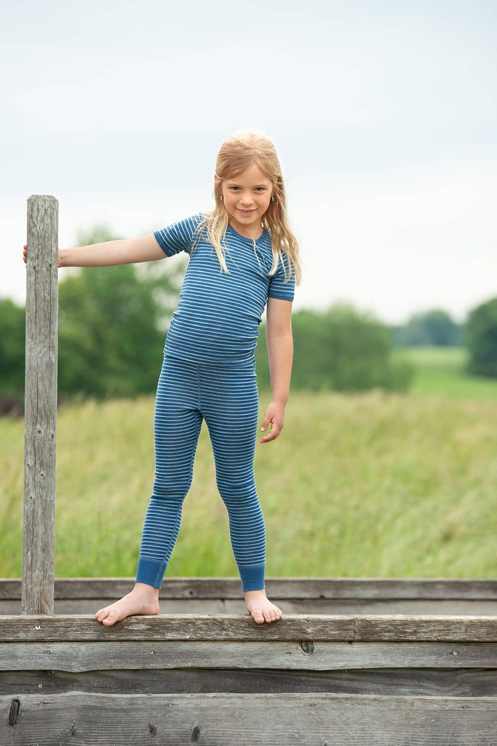 Geringelte Lange Wollunterhose blau/naturweiß Mädchen steht mit blau weiß geringelte lange Wollunterhose aus bio Wolle (kbT) und passendem Ringelshirt auf Holz
