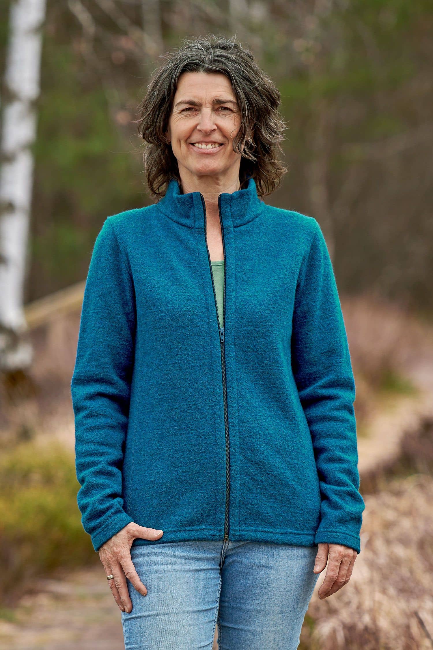 Frau steht im Wald und trägt Jeans, grünes Shirt und türkise Jacke