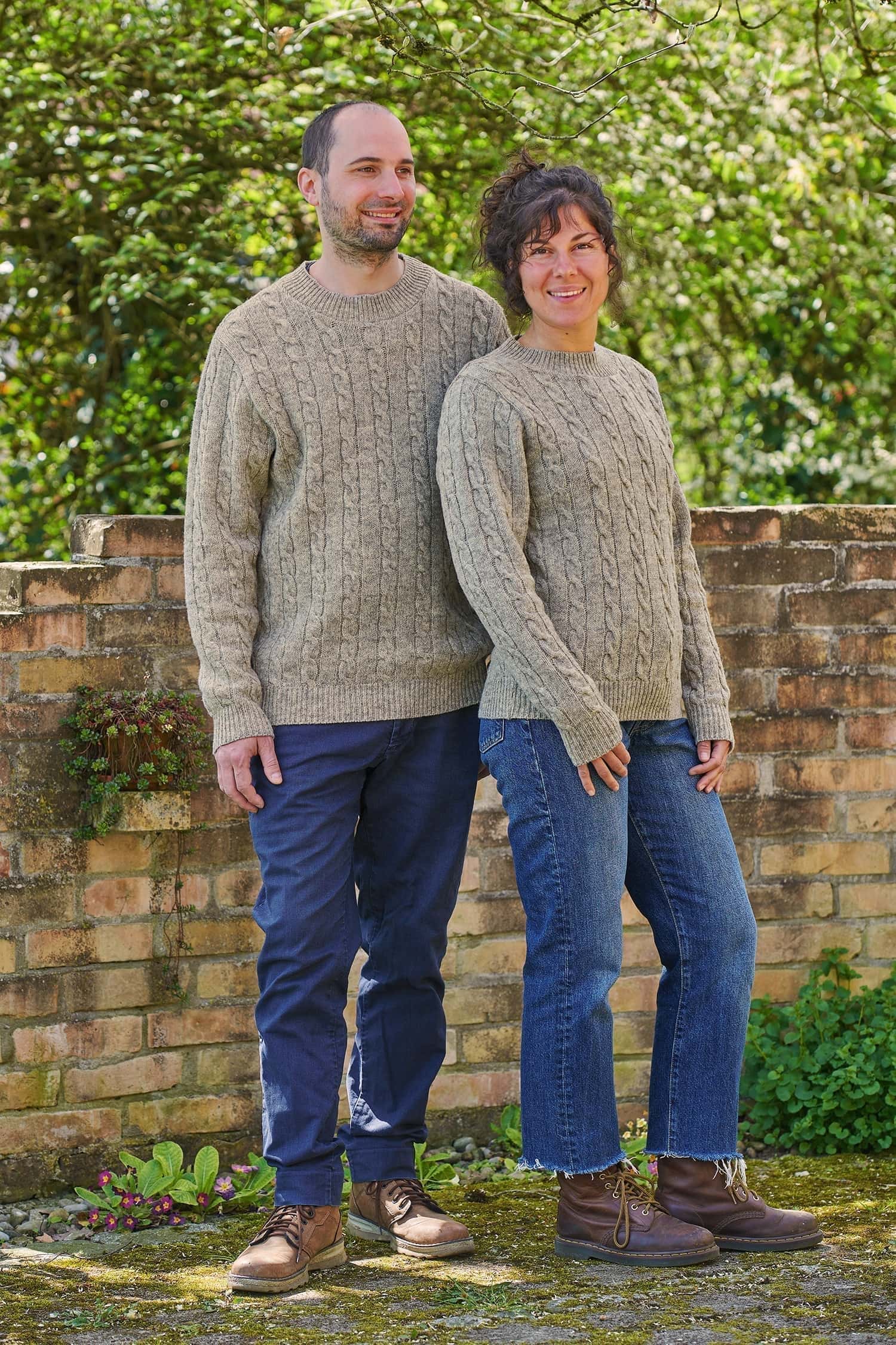 Mann und Frau tragen einen grauen Wollpullover vom Finkhof aus Bio-Wolle mit Zopfmuster und dazu Jeans und Stiefel