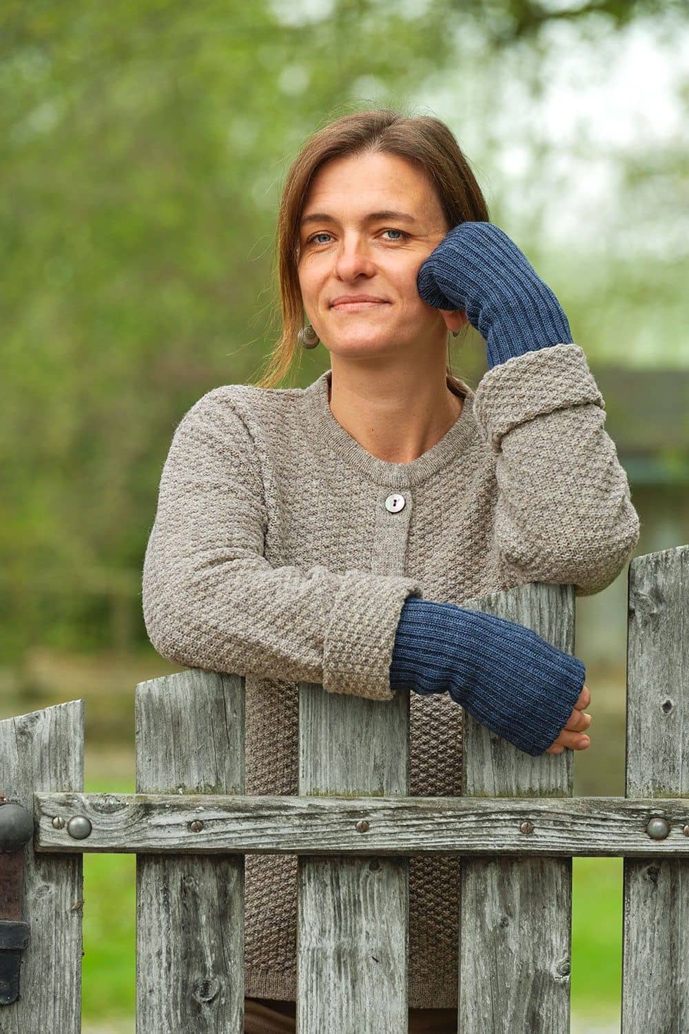 Frau mit grauer Strickjacke und jeansblauen Pulswärmern aus bio Wolle (kbT) vom Finkhof lehnt sich auf Zaun