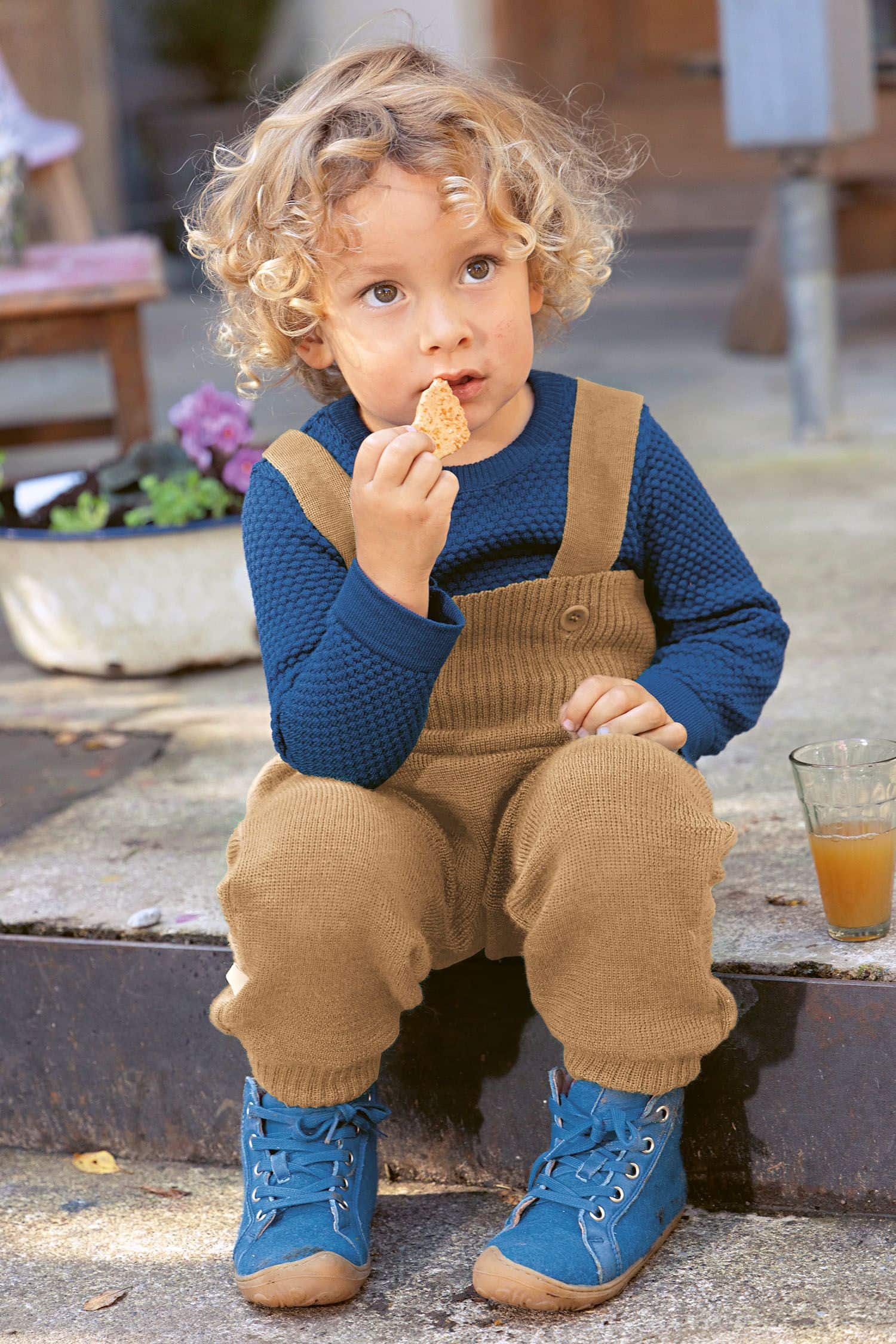 Kind sitzt auf Treppe und trägt braune Latzstrickhose aus bio Merinowolle (kbT), ein blaues Shirt und isst einen Keks