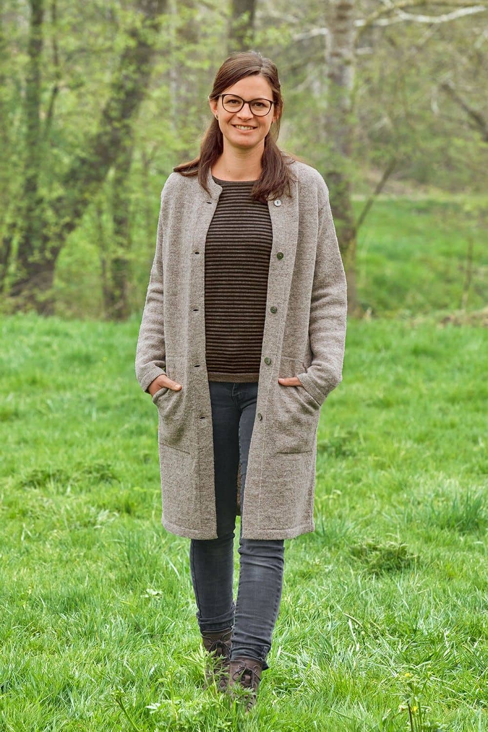 Frau auf Wiese trägt grauen offenen Finkhof Strickmantel aus bio Wolle (kbT) mit Mütze, Streifenshirt und Jeans