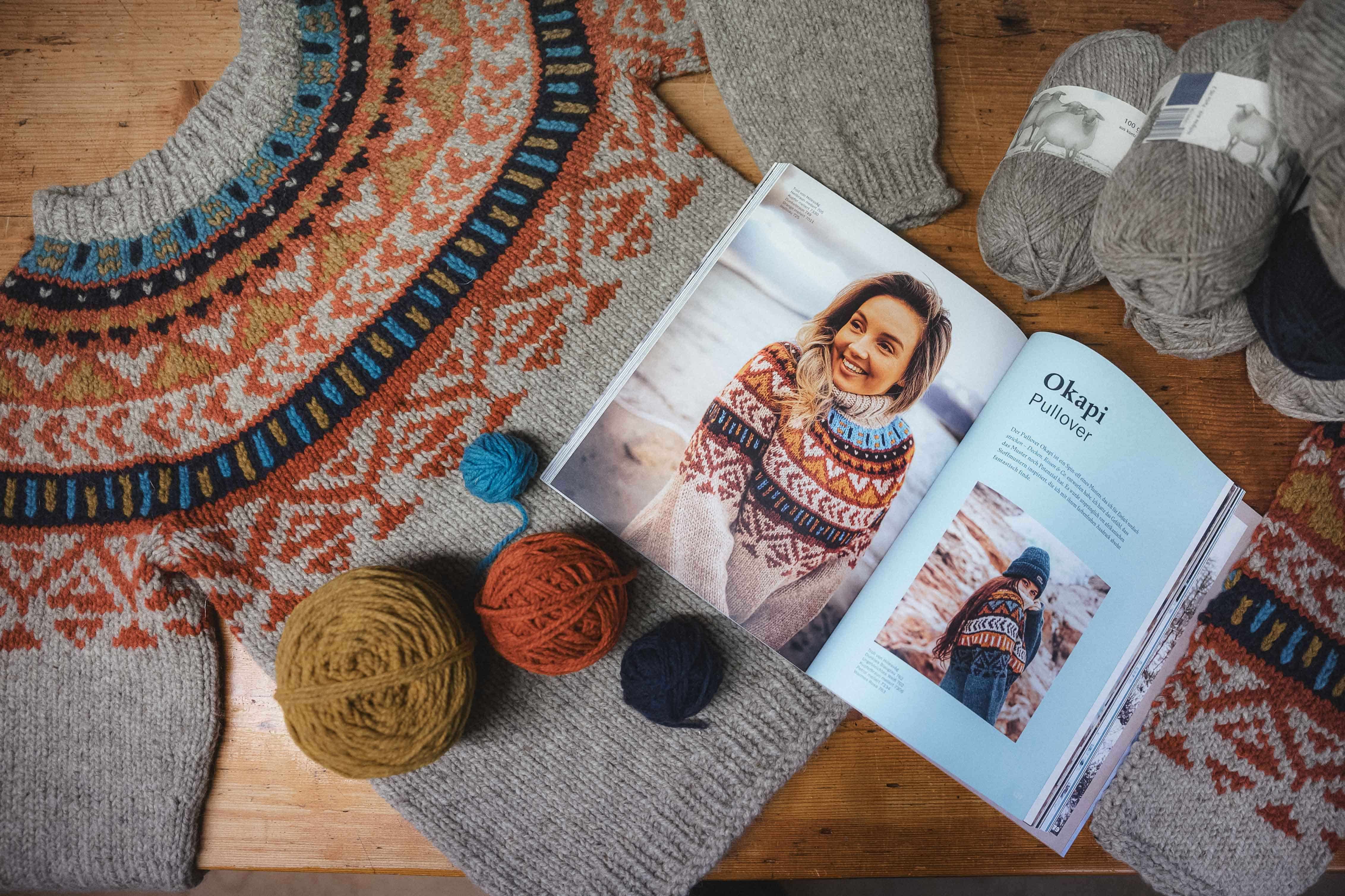 aufgeschlagenes Buch mit Strickanleitungen liegt auf grauem Norwegerpulli mit bunten Wollen