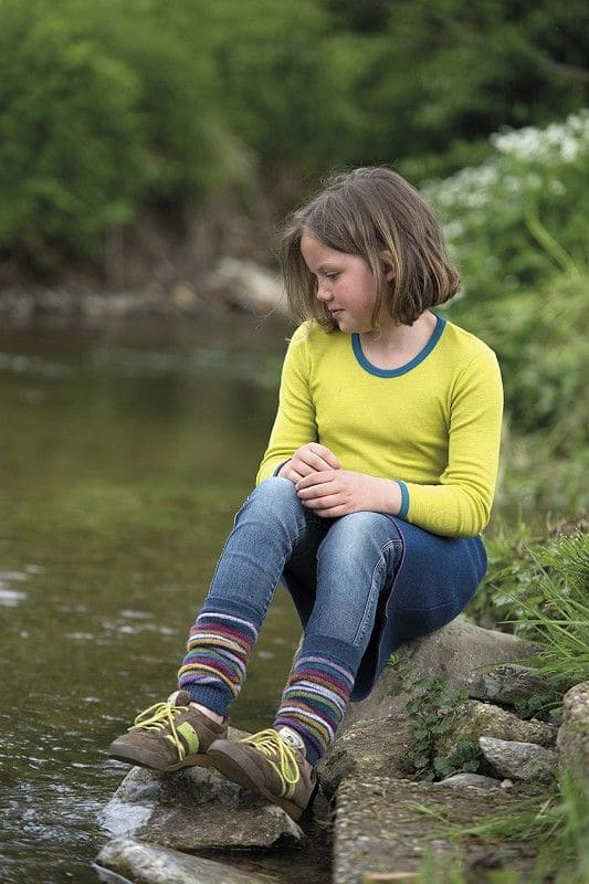 Kind sitzt auf Steinen an Bach mit grünem Shirt, Jeans und bunt gestrieften Beinstulpen aus bio Wolle (kbT)