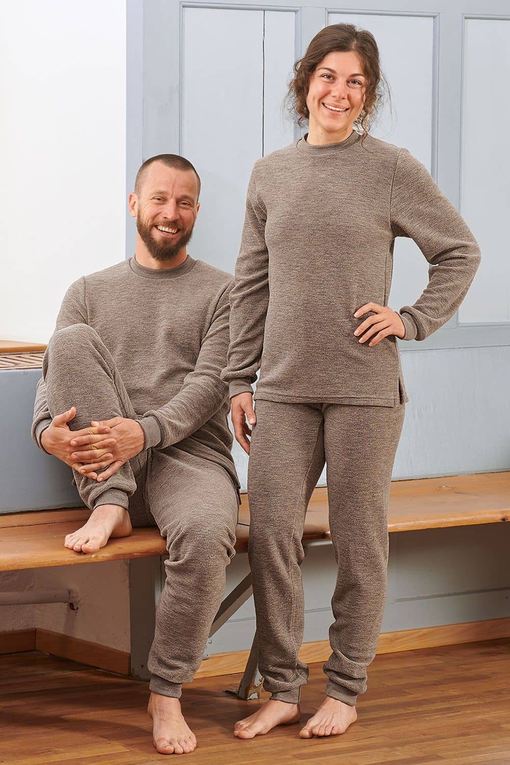 Mann sitzt auf Bank, Frau steht daneben und beide tragen walnussbraune Wollfrotte-Schlafanzüge aus bio Wolle (kbT)
