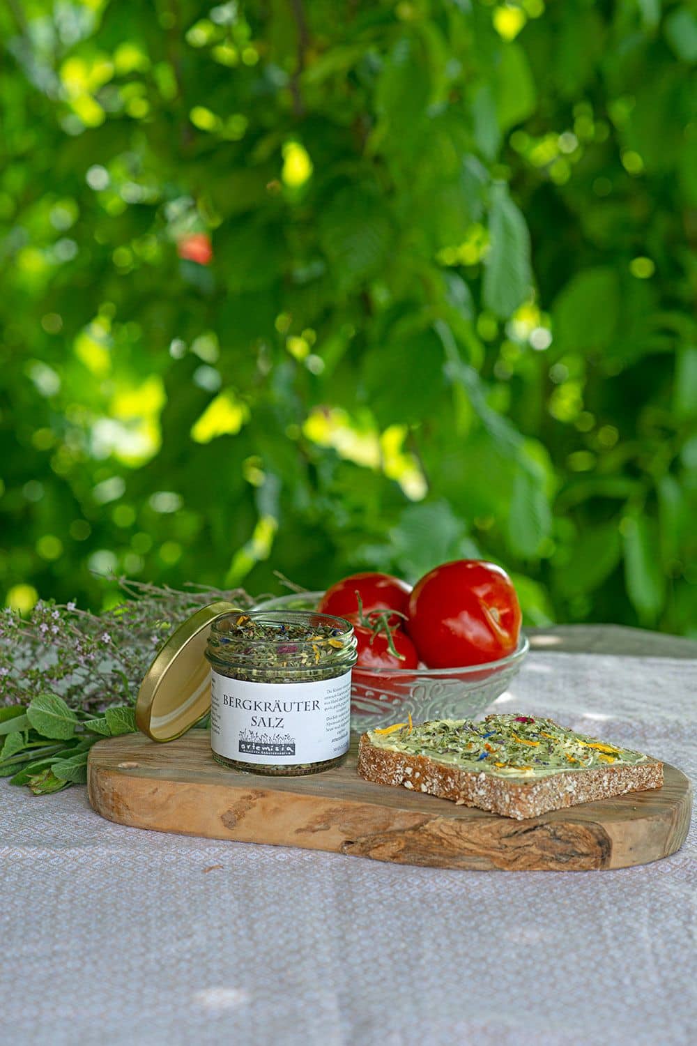 Bergkräutersalz Bergkräutersalz in geöffnetem 80g Glasdöschen mit Brot, Brettchen und Tomaten in Schale unter einem Baum