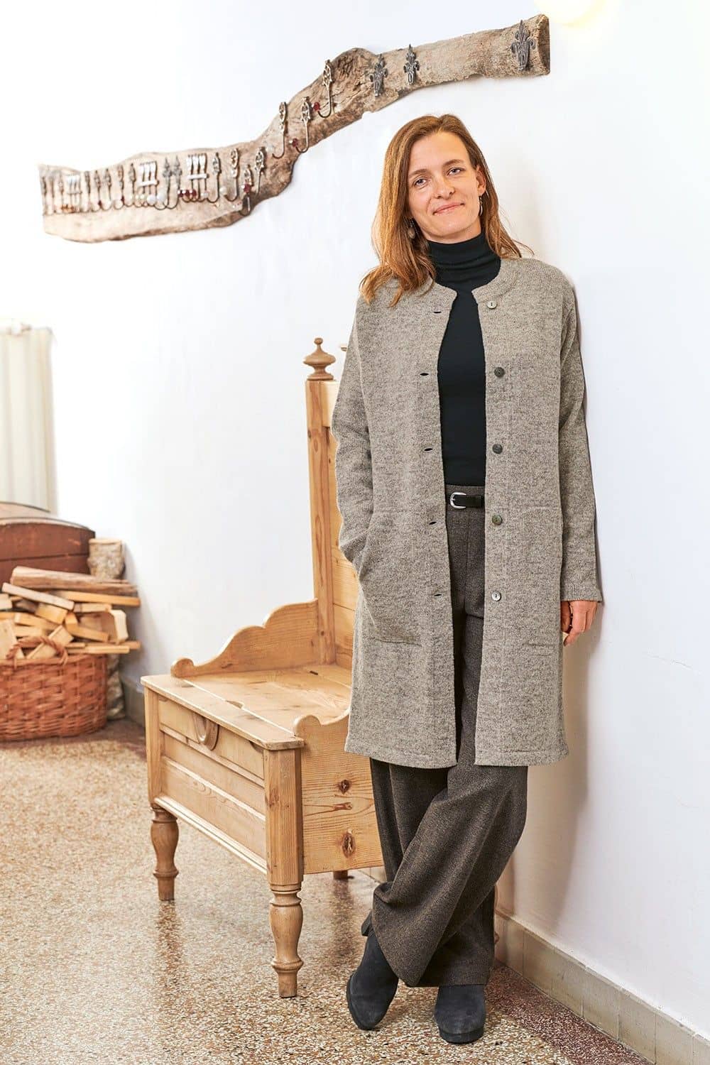 Frau lehnt an Wand und trägt Rolli, Wollhose und grauen offenen Finkhof Strickmantel aus bio Wolle (kbT) mit Knopfleiste