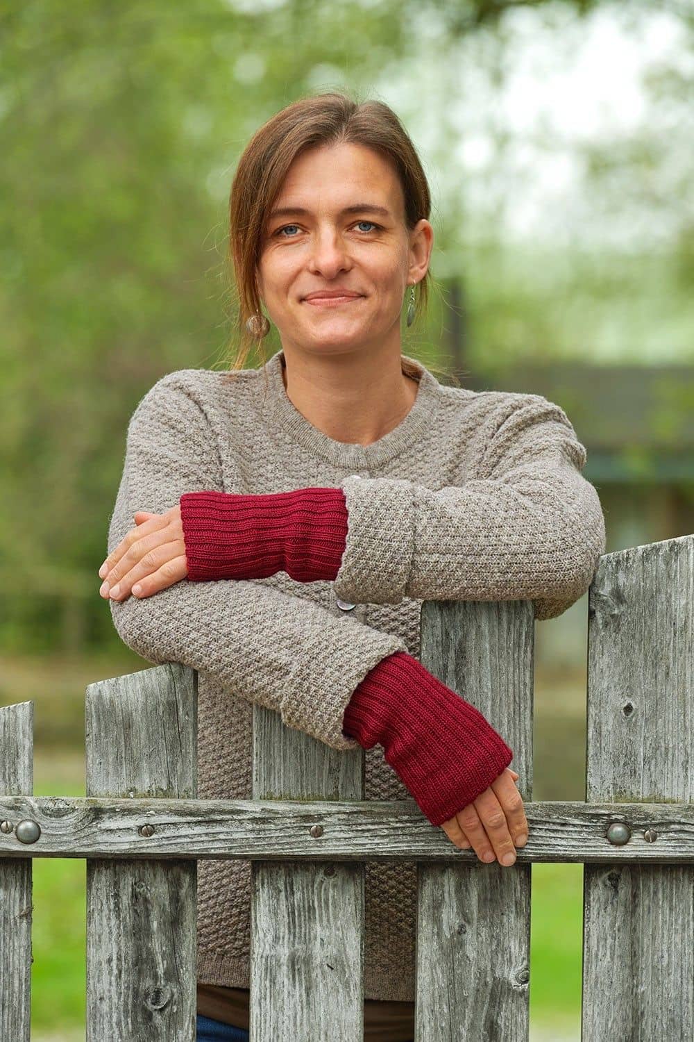 Frau mit grauer Strickjacke und roten Pulswärmern aus bio Wolle (kbT) vom Finkhof lehnt sich auf Zaun