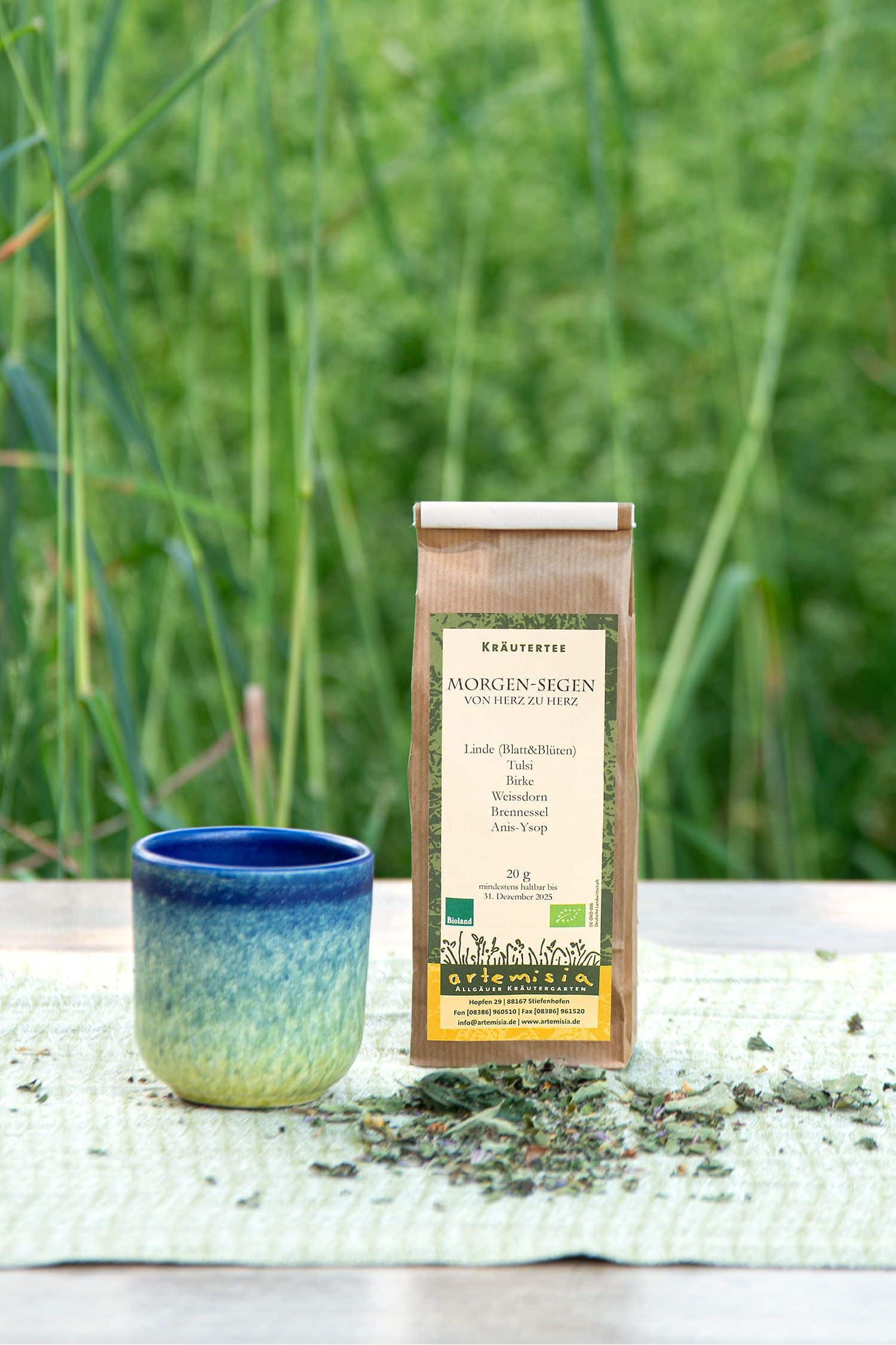 Morgen-Segen-Tee Morgen Segen Tee in 20g Packung neben Becher und getrockneten Kräutern vor einer Wiese