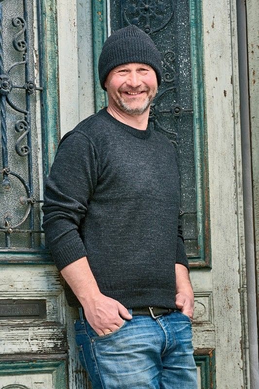 Mann lehnt sich mit dunkelgrauem Rundhalspullover aus bio Wolle (kbT) vom Finkhof und anthraziter Mütze an eine Tür