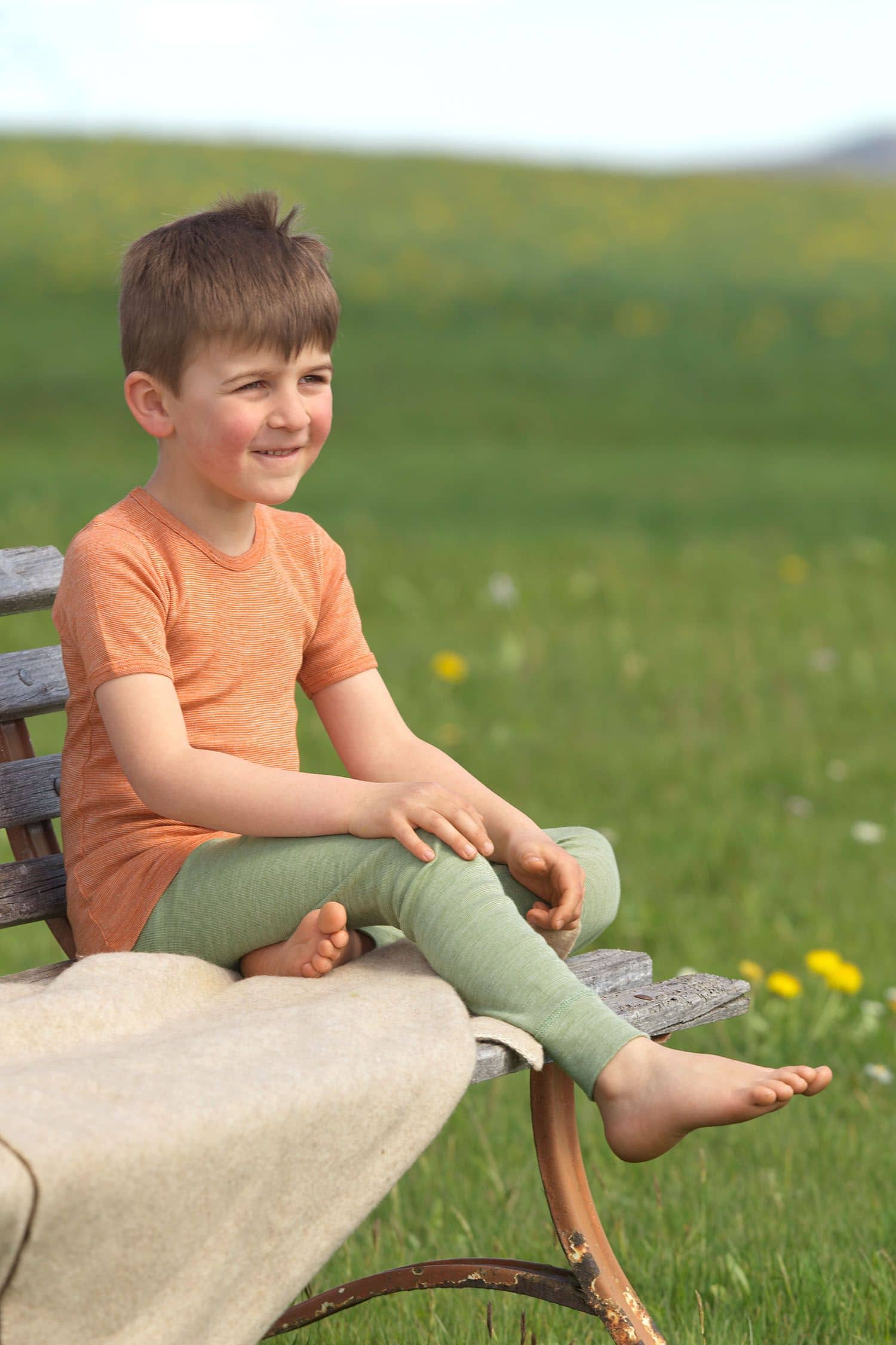 Junge sitzt auf Bank und trägt grüne lange Unterhose aus bio Wolle (kbT), bio Baumwolle (kbA) und Seide mit orangem Shirt