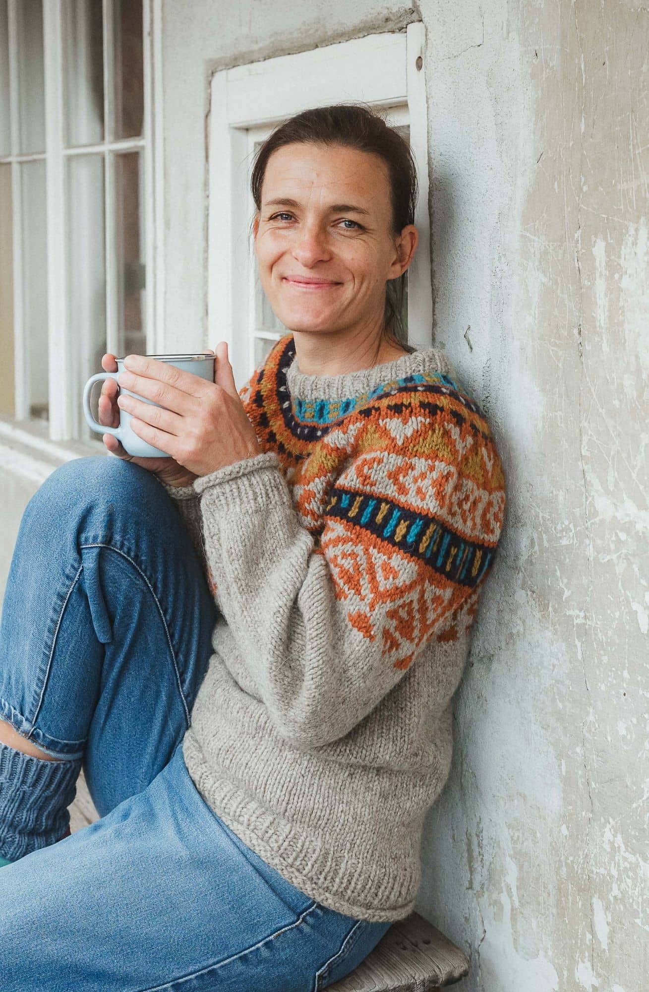 Frau sitzt auf einer Bank mit einer Tasse in der Hand und trägt einen grau orangegemusterten Norwegerpulli