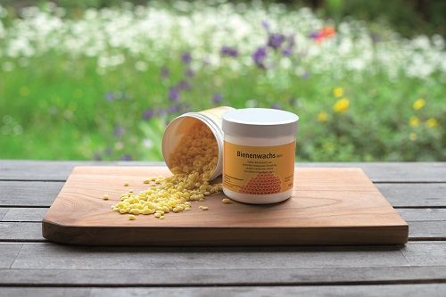 weiße Döschen mit Bienenwachs stehen auf Holzbrettchen, eine Dose ist geöffnet umgekippt und im Hintergrund ist eine Wiese