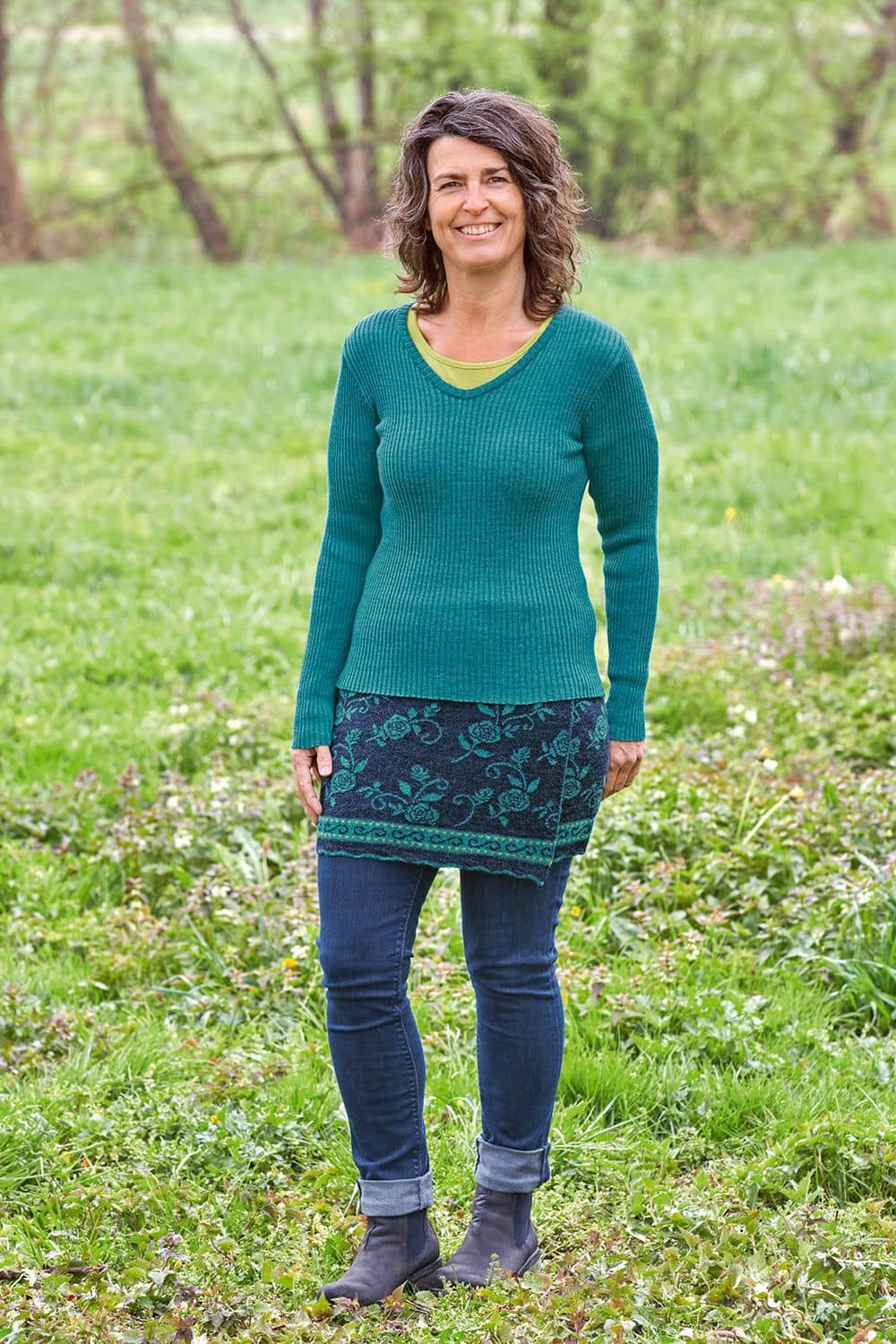 Cacheur Rose Frau steht auf Wiese und trägt blau grauen floralgemusterten Cacheur aus bio Wolle (kbT), türkises Rippshirt und Jeans