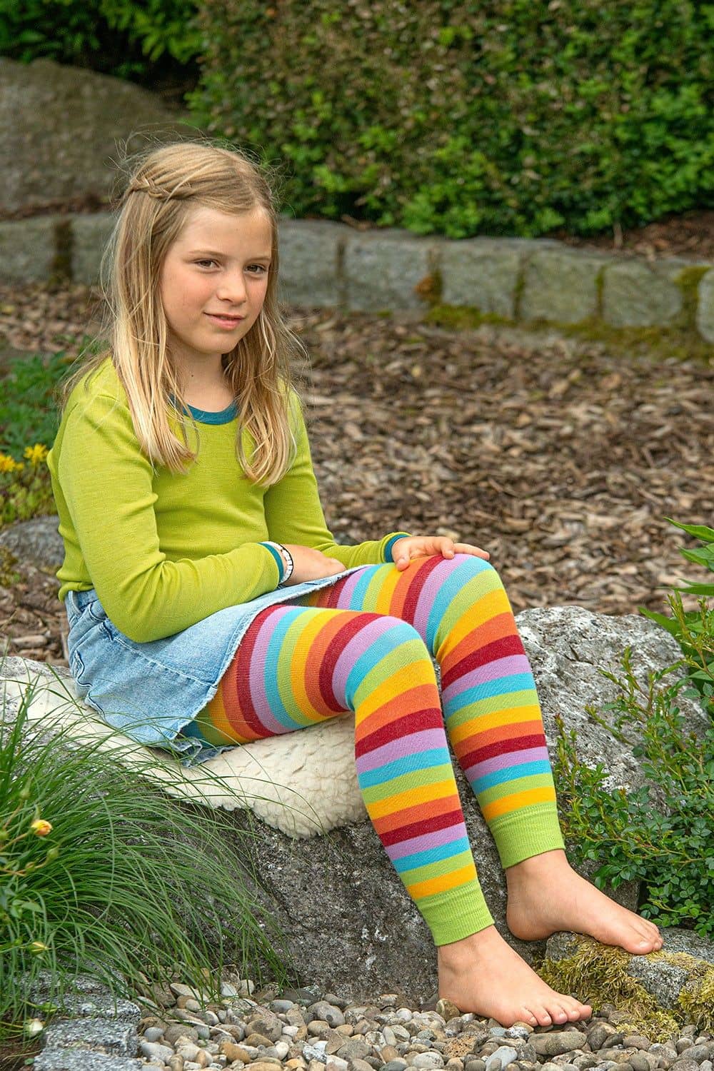 Kind sitzt auf Stein und trägt Jeansrock, grünes Shirt und bunte gestreifte Leggings aus bio Baumwolle (kbA) und Elasthan