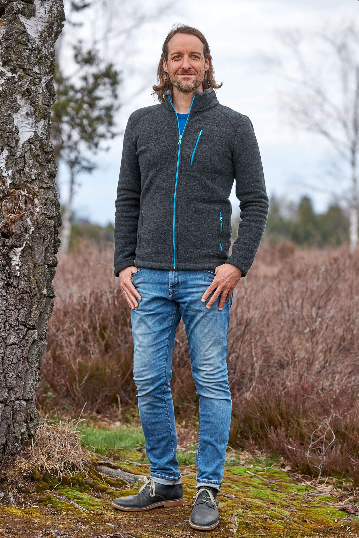 Mann steht neben Baum und trägt dunkelgraue Wollfleece Jacke aus bio Wolle (kbT) und eine Jeans