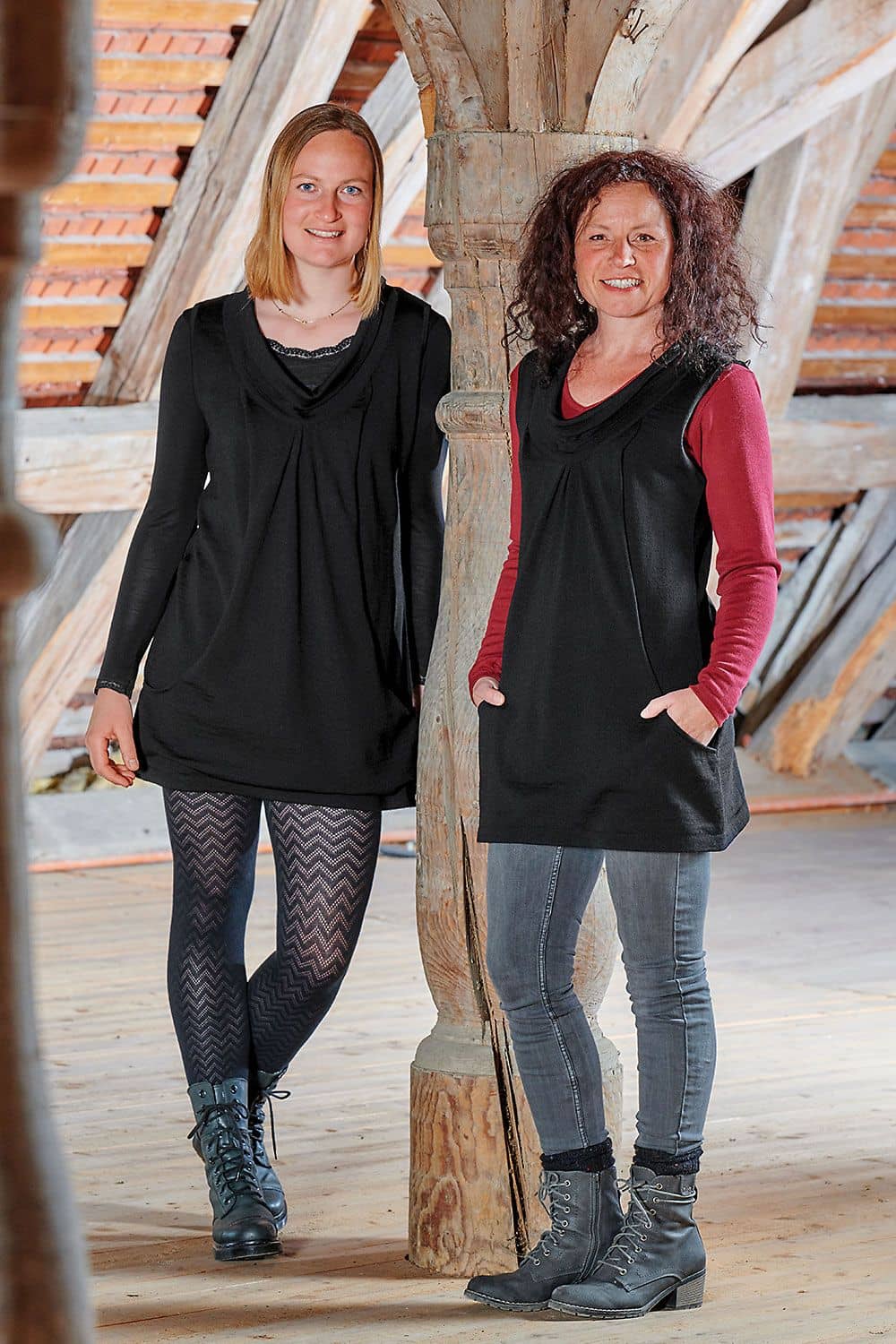 Frauen lehnen an Holzbalken und tragen schwarze Tunikas mit Taschen. Eine kombiniert sie mit schwarzer Ajourleggings