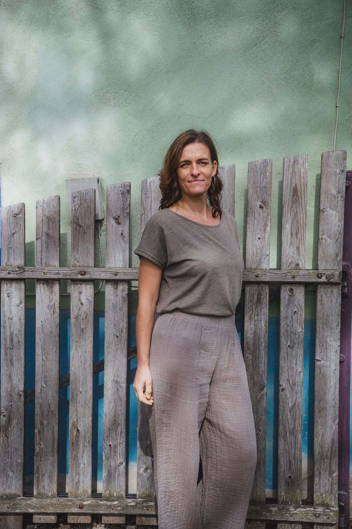 Frau steht vor Zaun und trägt ein Shirt und eine luftige braune Hose aus Bio-Baumwoll-Musselin (kbA) vom Finkhof