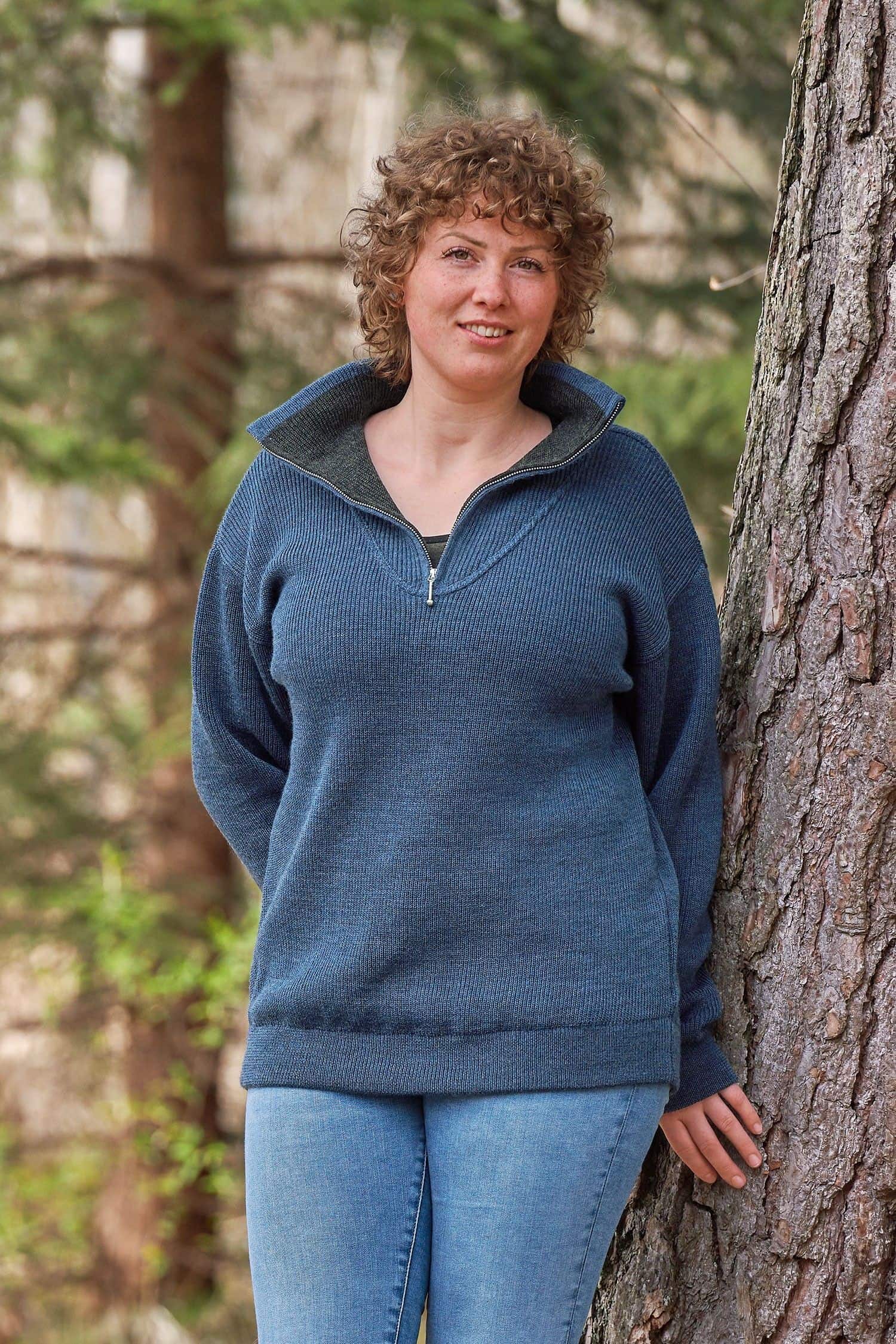 Frau steht neben Baum und trägt geöffneten rauchblauen Finkhof Troyer mit Zipper aus bio Wolle (kbT)