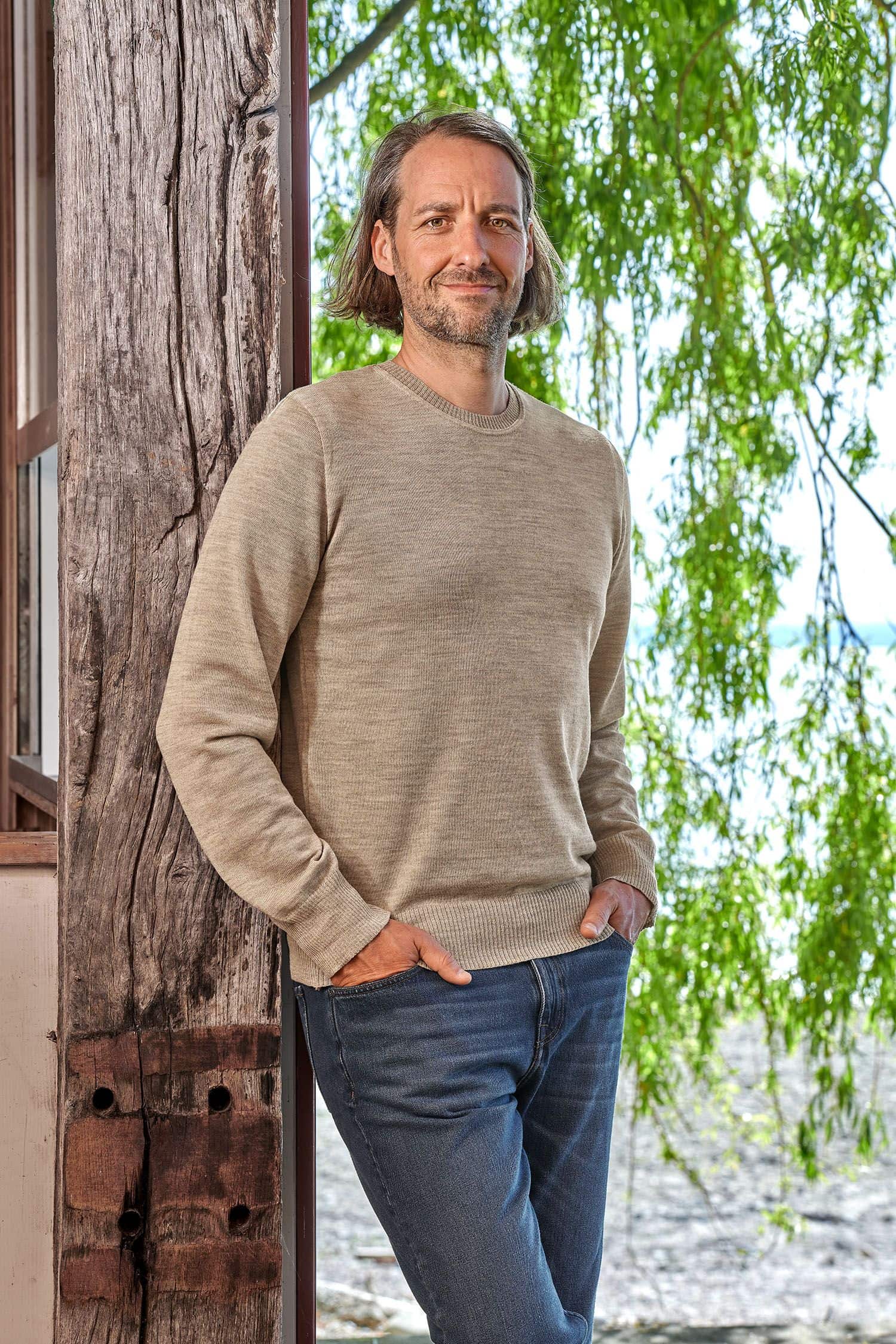 Herren-Wollpullover fein Mann mit feinem beigen Wollpullover aus bio Merinowolle (kbT) vom Finkhof lehnt an einem Balken