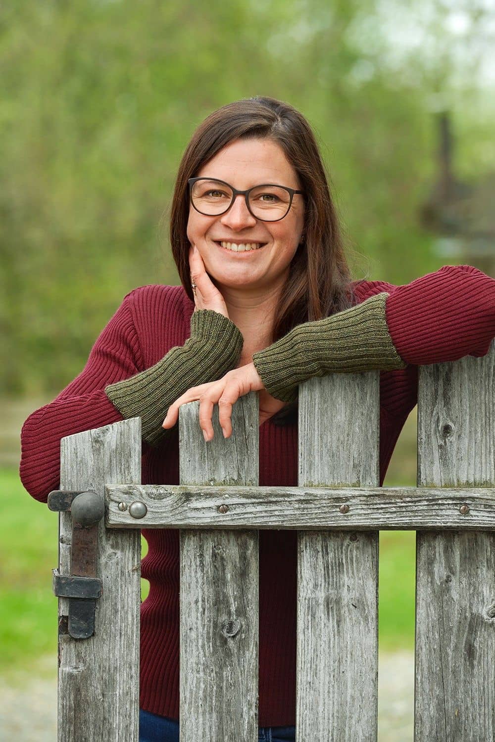 Frau mit rotem Rippshirt und olivegrünen Pulswärmern aus bio Wolle (kbT) vom Finkhof lehnt sich auf Zaun