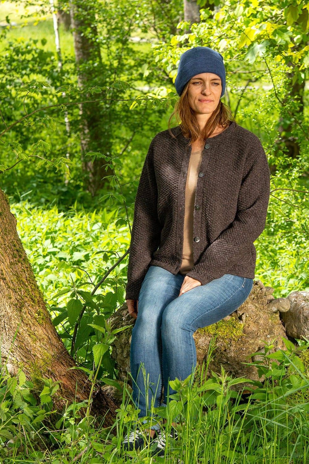 Frau sitzt auf Stein und trägt eine blaue leichte Mütze aus bio Wolle (kbT) und bio Baumwolle (kbA) und eine Strickjacke