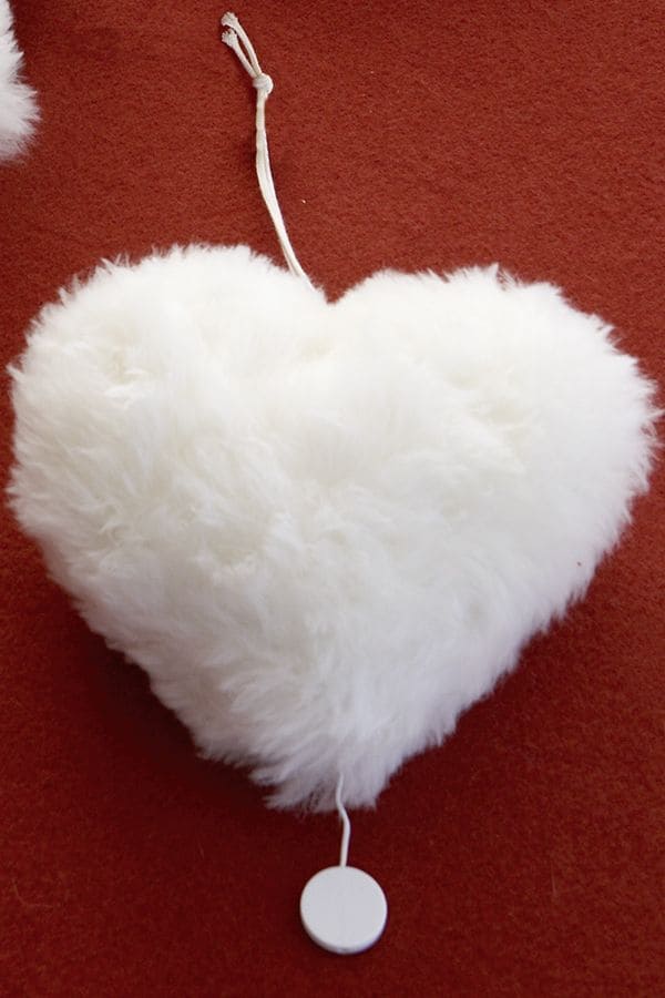 Nahaufnahme von weißer Finhof Spieluhr in Herzenform aus bio Wolle (kbT) und Fell liegt auf rotem Untergrund