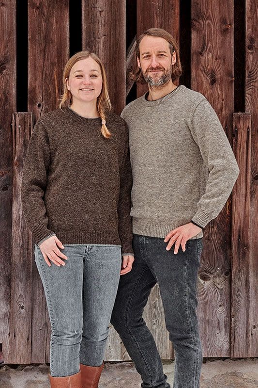 Finkhof-Pulli Mann und Frau vor Holztor tragen Finkhof Pullis aus bio Wolle (kbT) in braun und grau mit dunkler Jeans