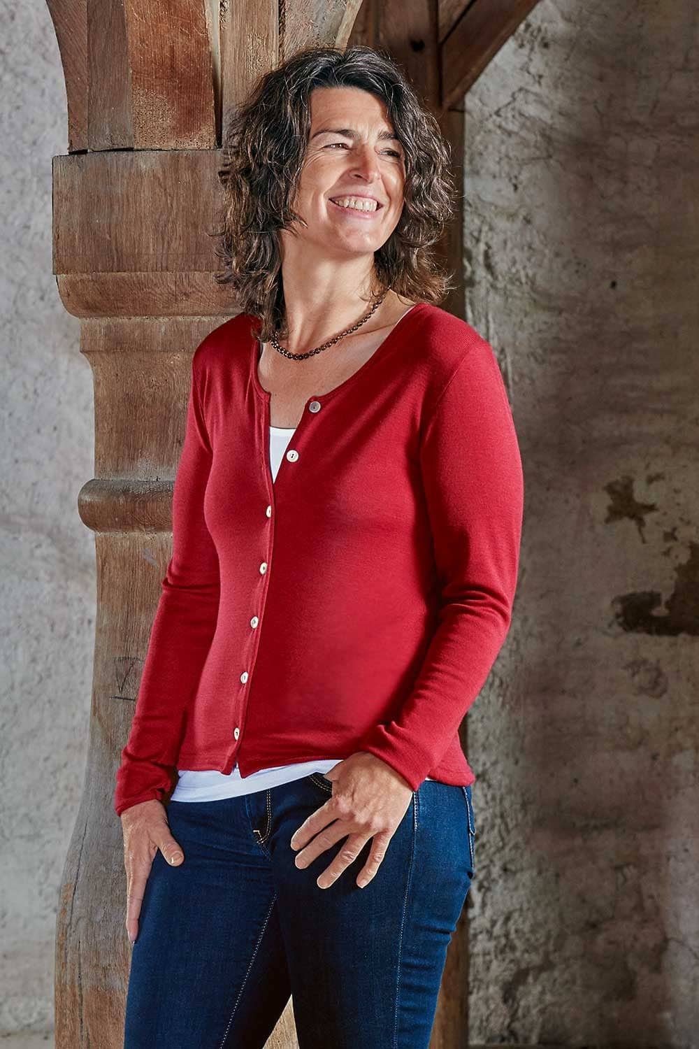 Frau lehnt mit rotem Cardigan aus bio Wolle (kbT) und Seide, weißem Top und Jeans an Balken