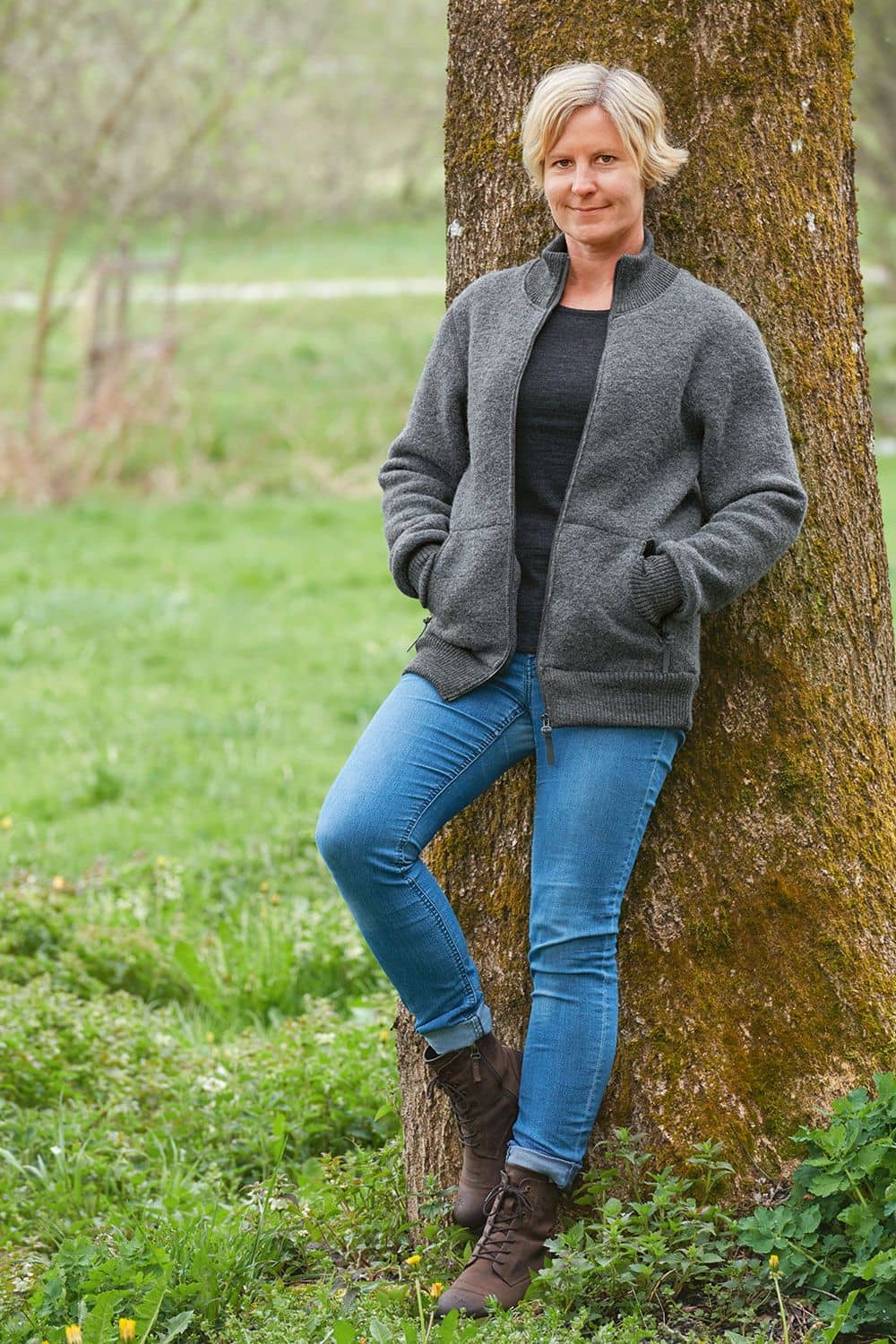 Frau an Baumstamm gelehnt trägt geöffnete graue Finkhof Jacke Toni aus bio Wolle (kbT) mit Jeans und Stiefeln