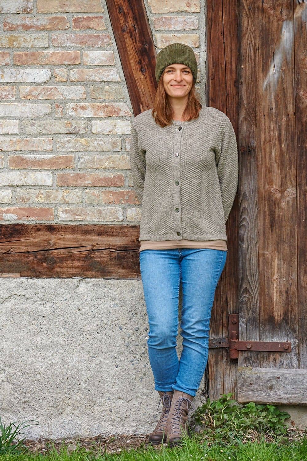 Frau mit Strickajcke und olivegrüner Wollmütze aus bio Wolle (kbT) vom Finkhof lehnt sich an Wand