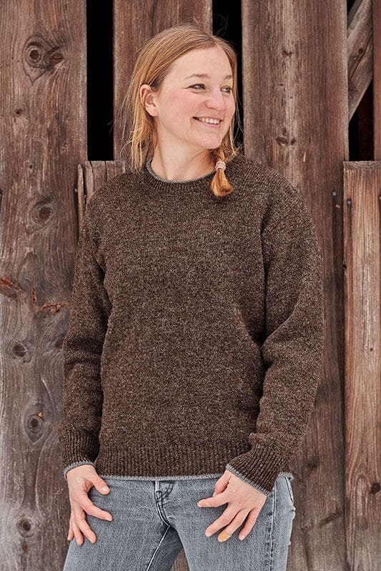 Frau mit blondem Zopf vor Holzwand trägt braunen Finkhof Pulli aus bio Wolle (kbT) mit Rundhalsausschnitt