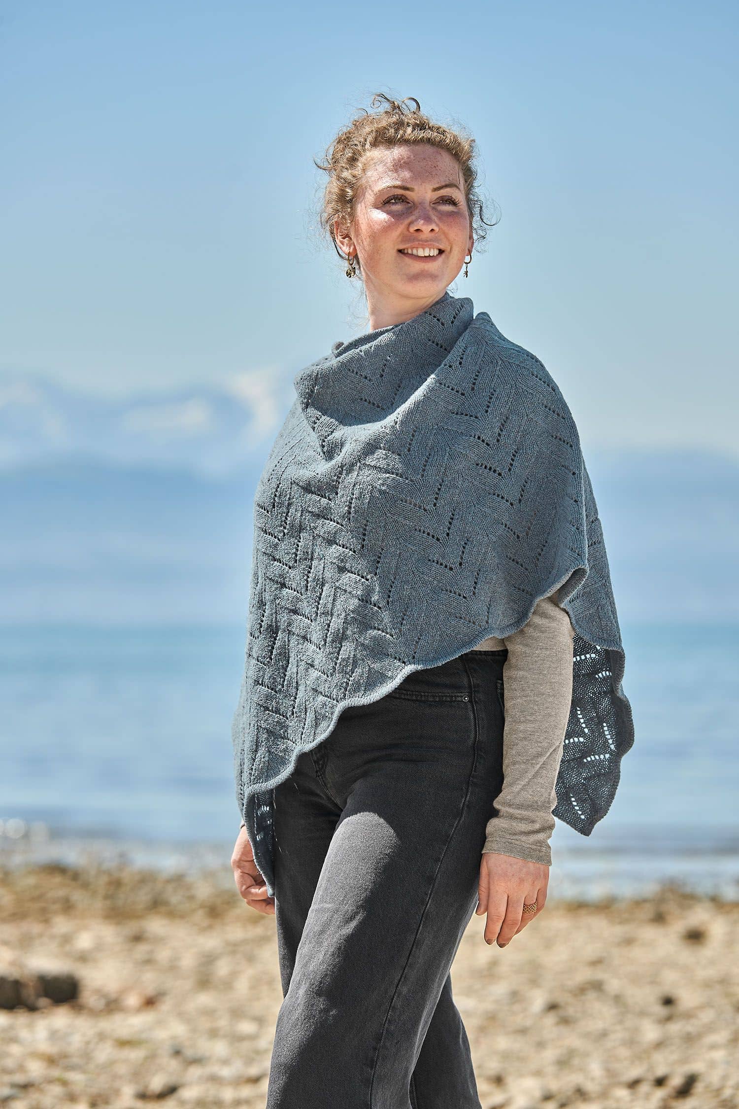 Frau trägt eine schwarze Jeans und einen rauchblauen Poncho mit Ajour Muster vom Finkhof aus bio Merinowolle (kbT)