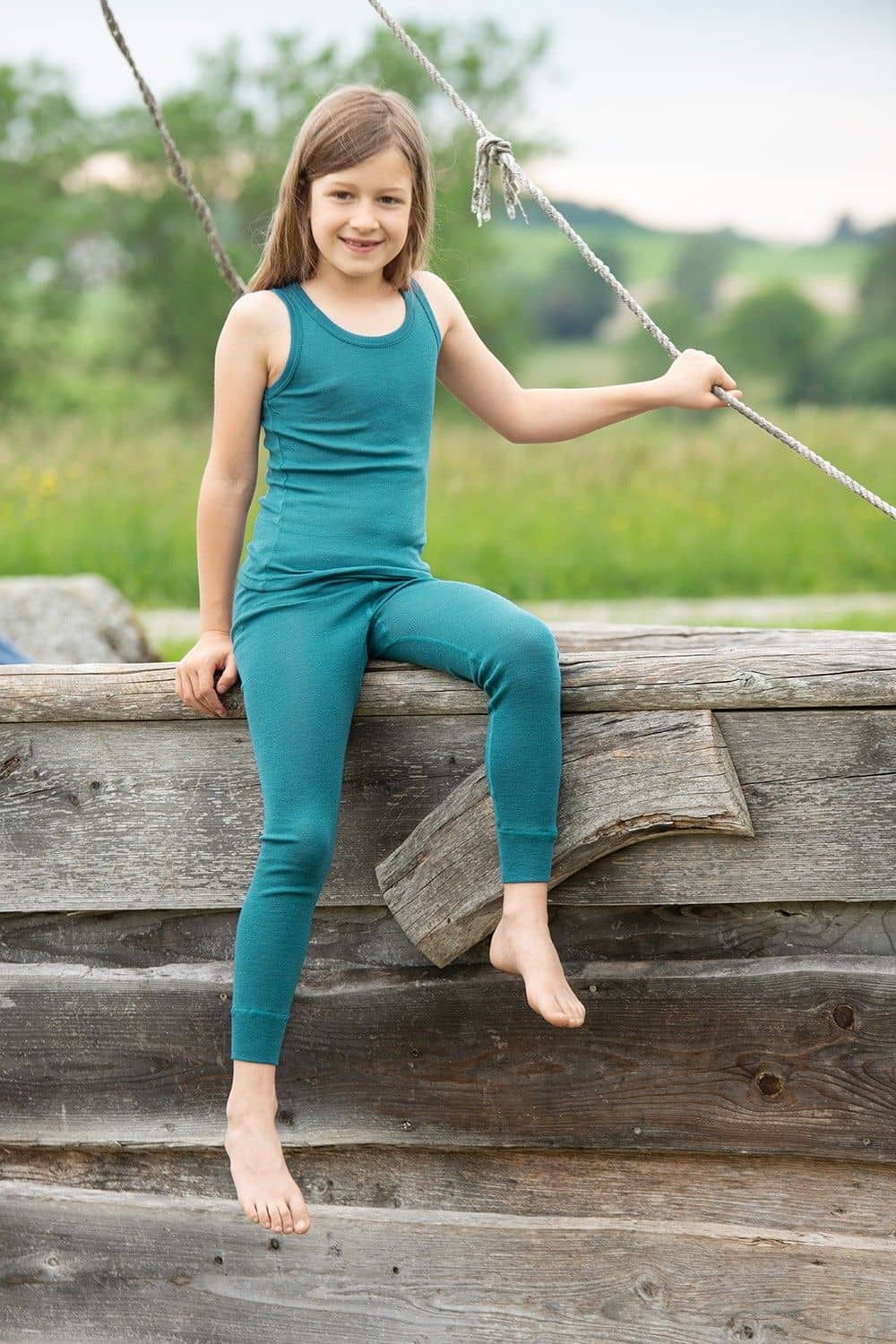 Kind sitzt auf Holzschiff und trägt eine türkise lange Unterhose und ein Unterhemd aus bio Wolle (kbT) und Seide