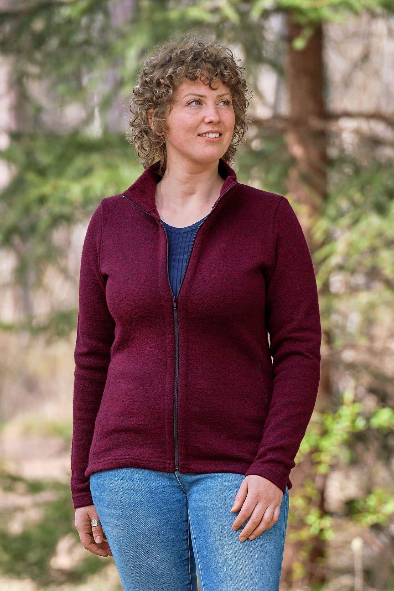 Frau steht im Wald und trägt Jeans, blaues Shirt und beerefarbene gewalkte Jacke
