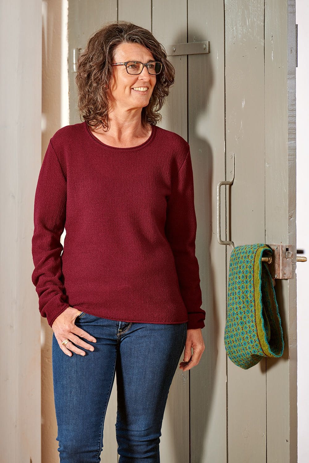 Frau trägt bordeauxroter Wollpulli aus bio Wolle (kbT) vom Finkhof, an Türklinke hängt grün gemusterter Schal