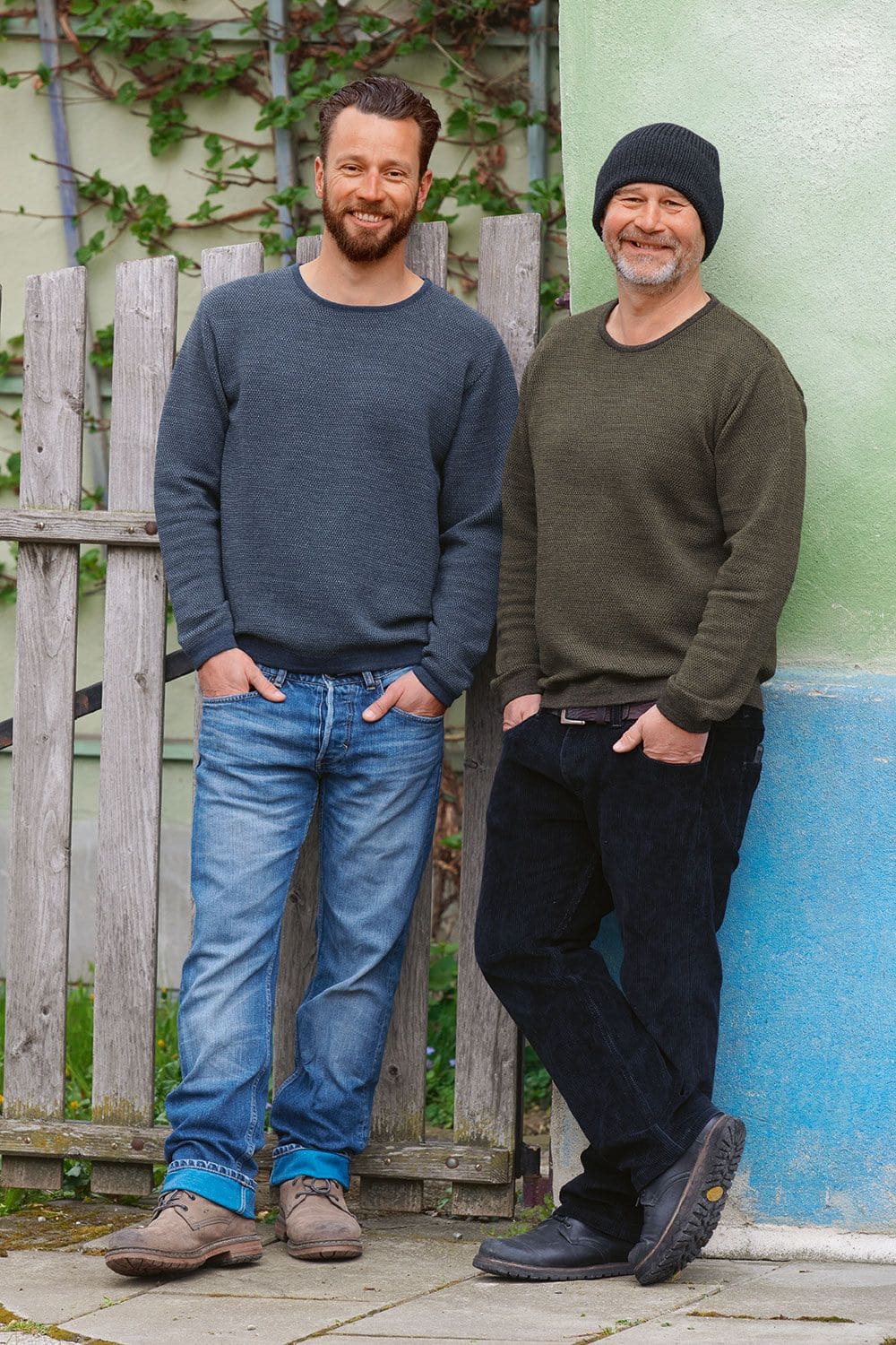 Melangepullover Männer an Wand gelehnt tragen blauer und grüner Finkhof Melangepullover aus bio Wolle (kbT) mit Jeans und Stiefeln